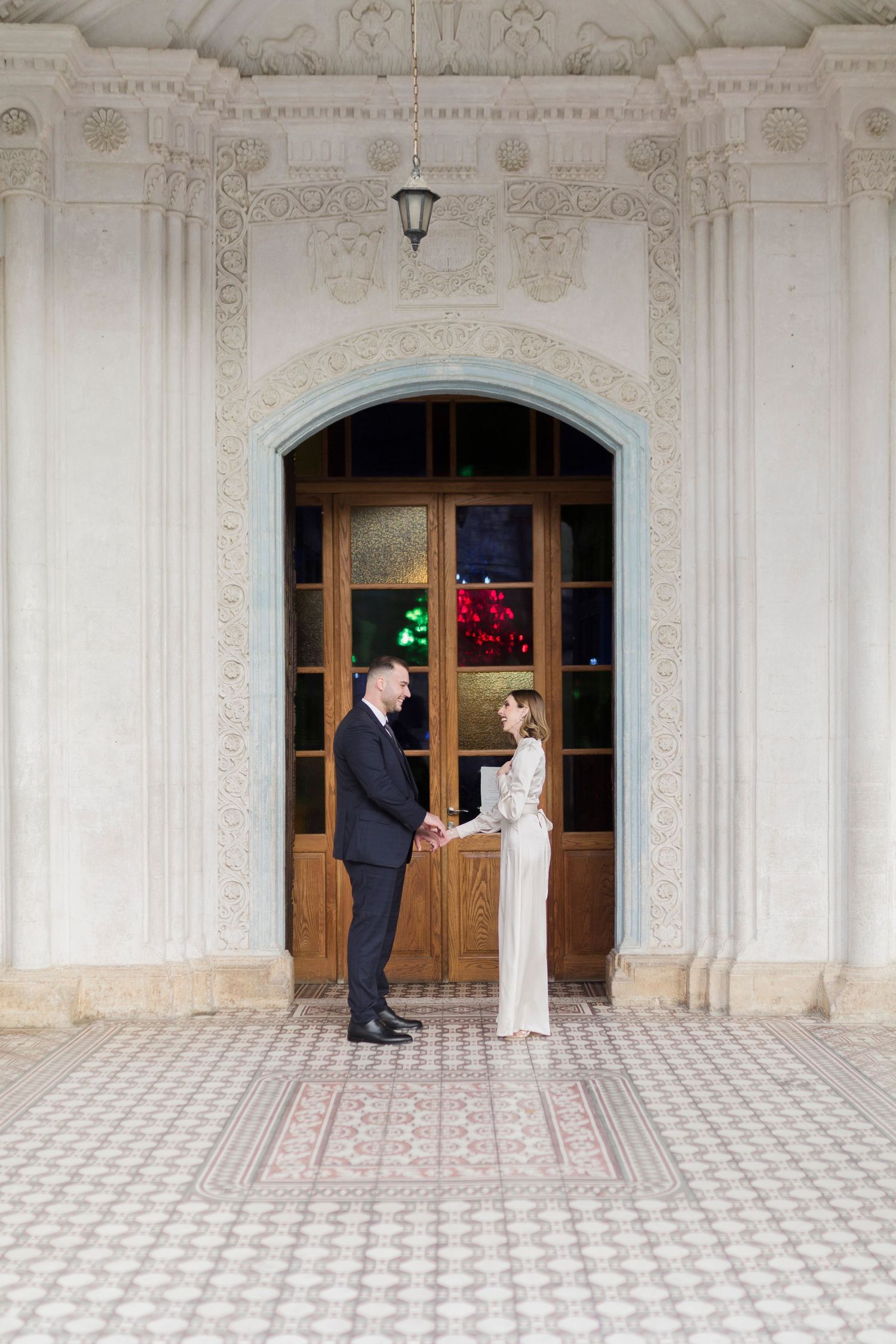 Elegant couple holding hands in front of ornate white building with wooden arched doors during wedding ceremony.