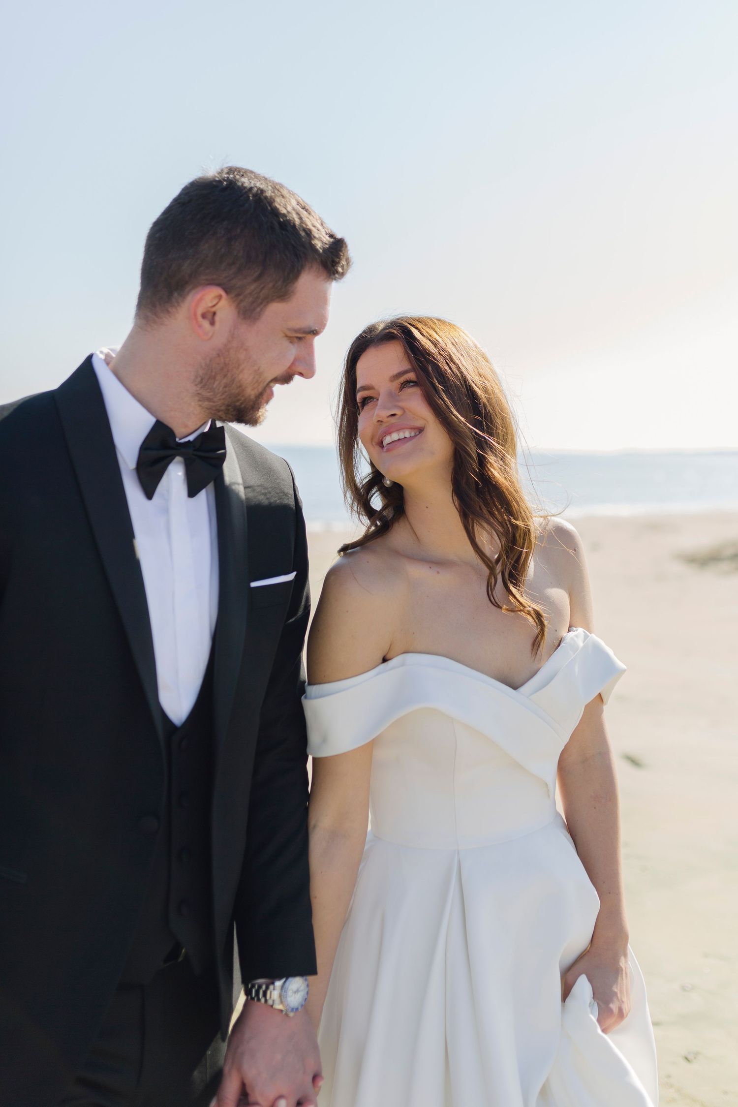 A smiling bride in an off-shoulder white gown holds hands with a groom in a black tuxedo on a sunny beach.