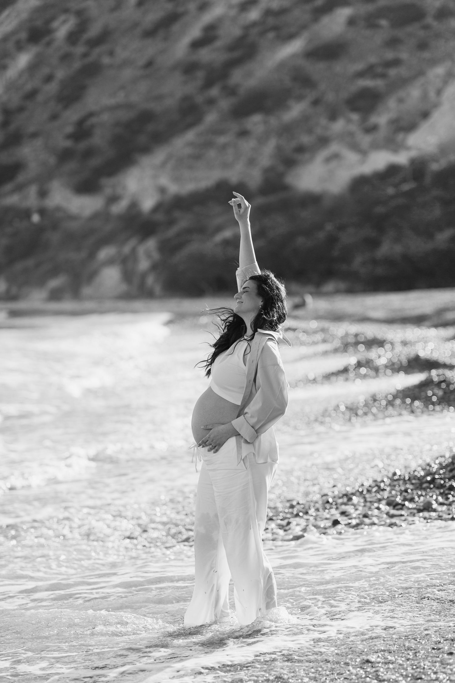 Black and white photo of a pregnant woman in white standing in ocean waves, cradling her belly on a pebble beach.