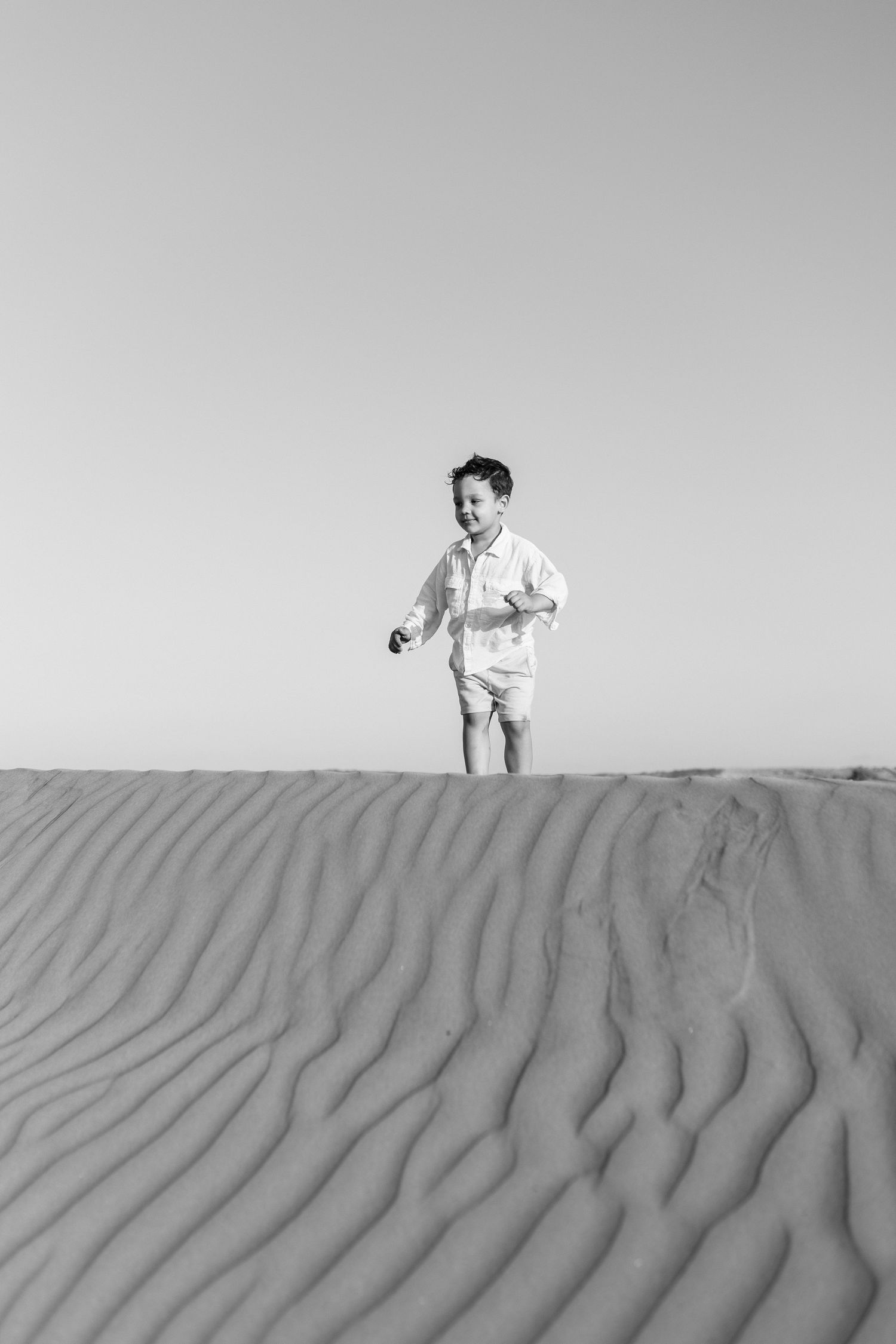 Black and white photo of a young boy running joyfully across rippled sand dunes in the desert.
