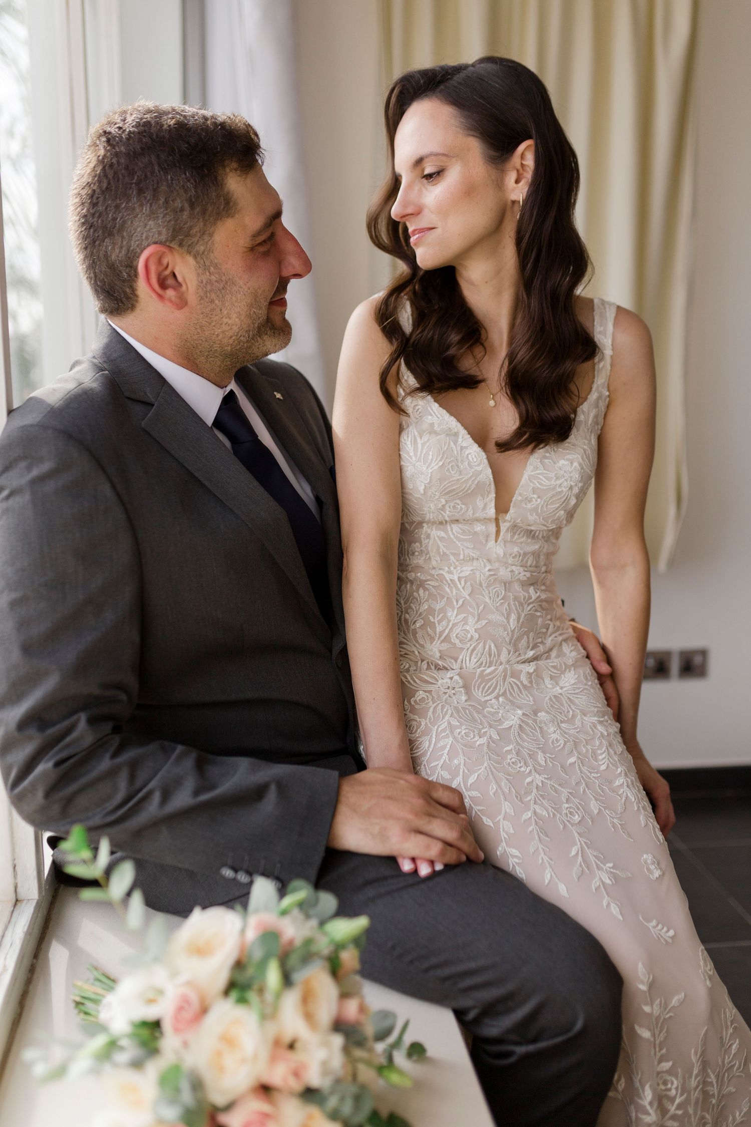 Bride in lace gown and groom in gray suit share an intimate moment seated by a window with a floral bouquet.