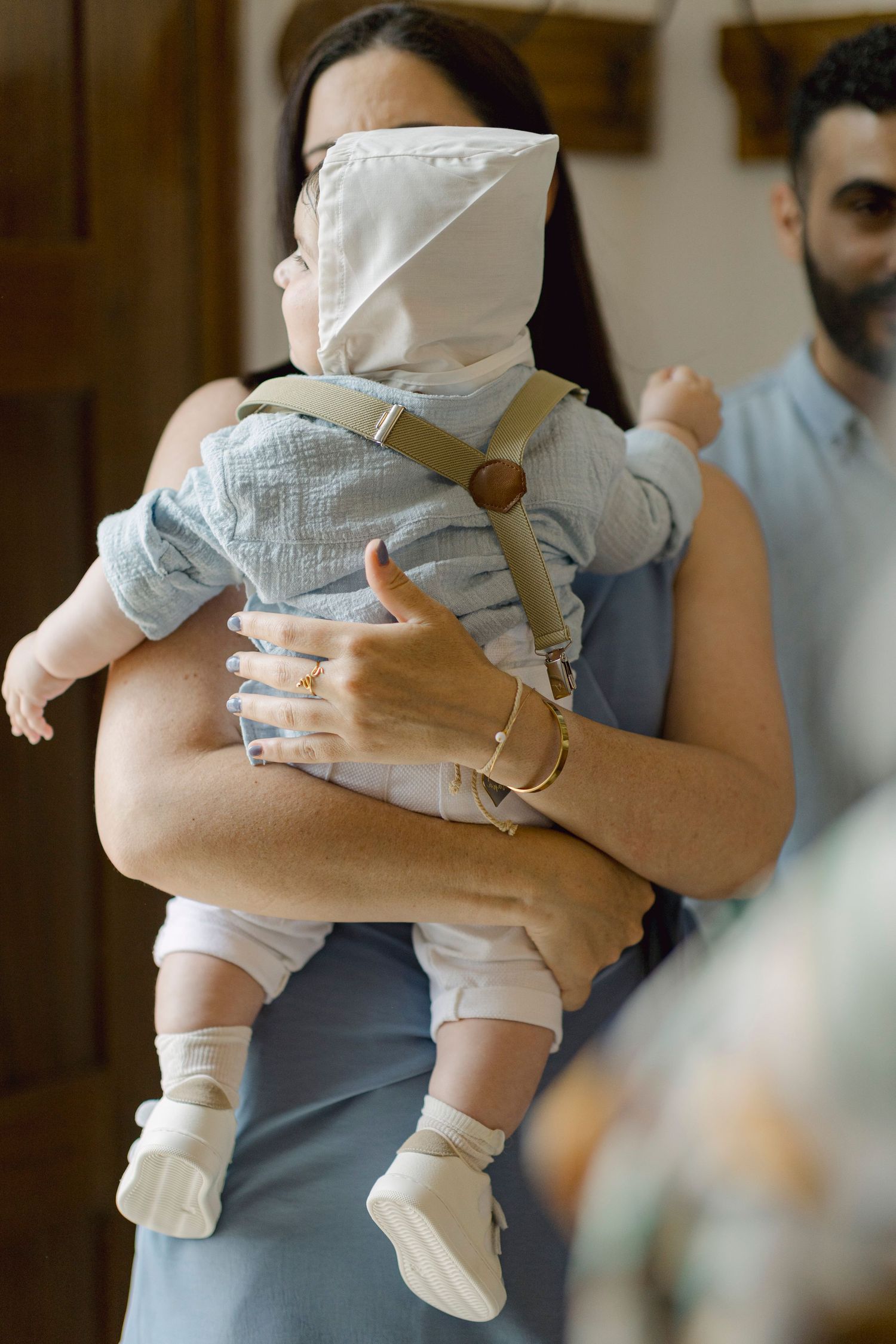A woman in a blue dress holds a baby wearing a white bonnet and suspenders at an indoor gathering.