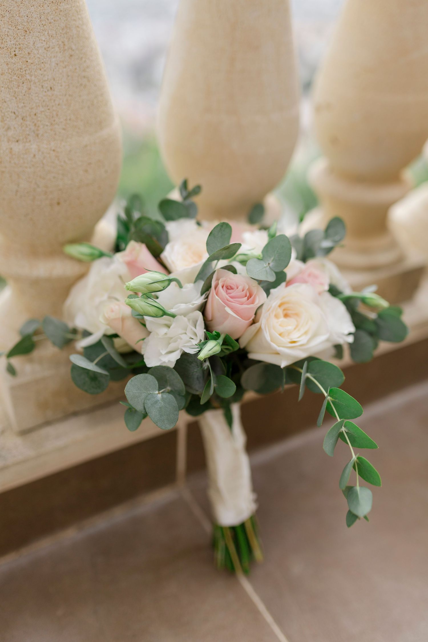 Wedding bouquet of blush pink and white roses with eucalyptus resting against stone balustrade.