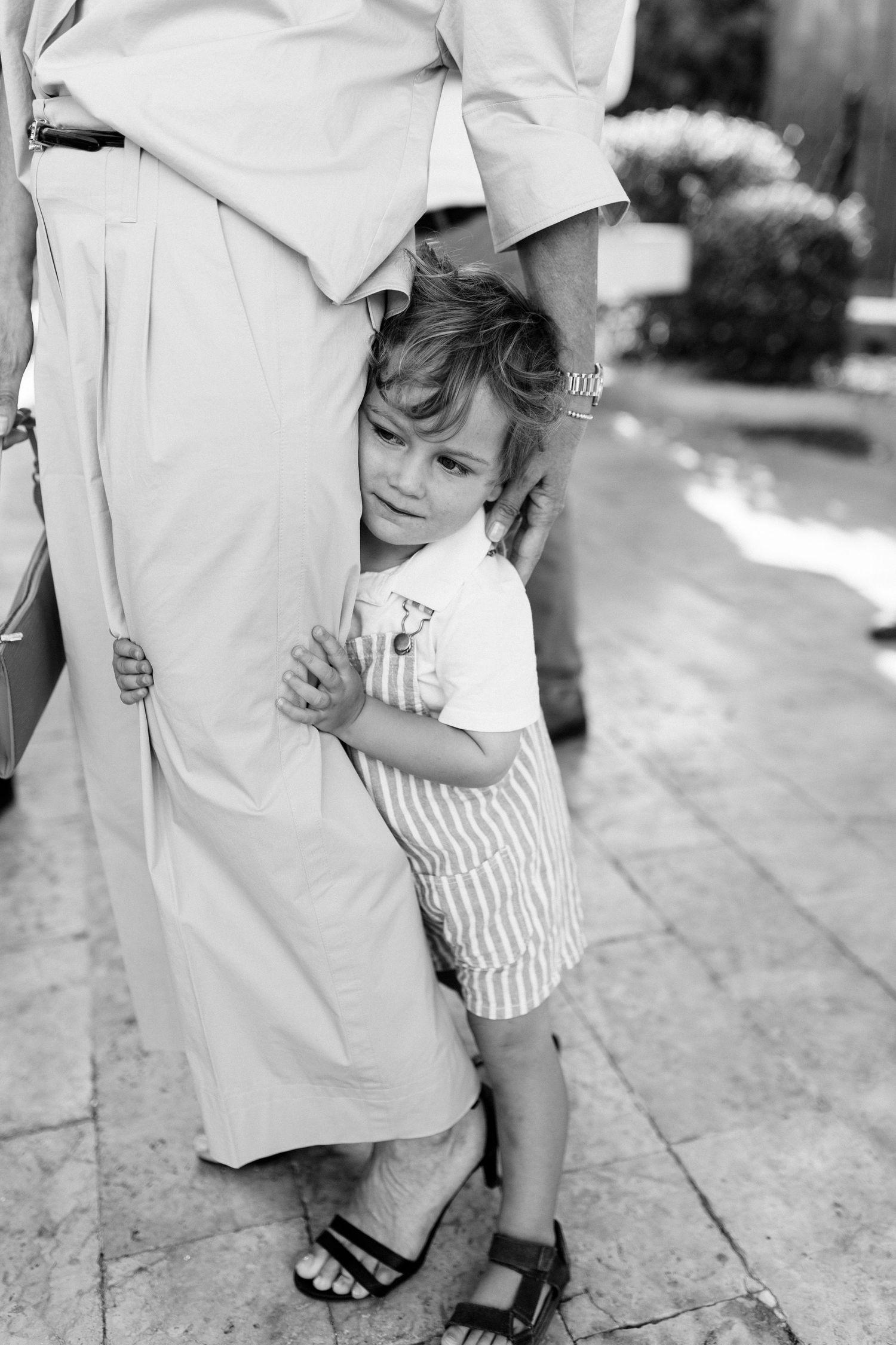 Black and white photo of a young child clinging to an adult's leg outdoors, wearing striped shorts and sandals.