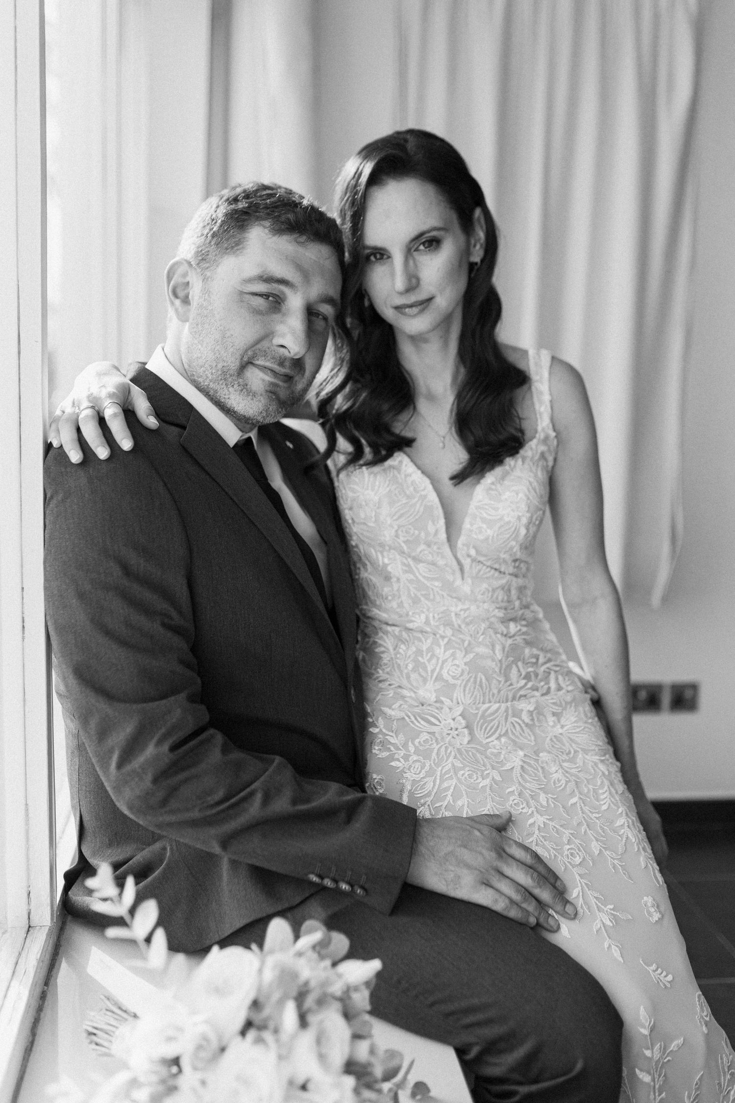 Intimate black and white portrait of bride and groom posing together by window in elegant wedding attire.