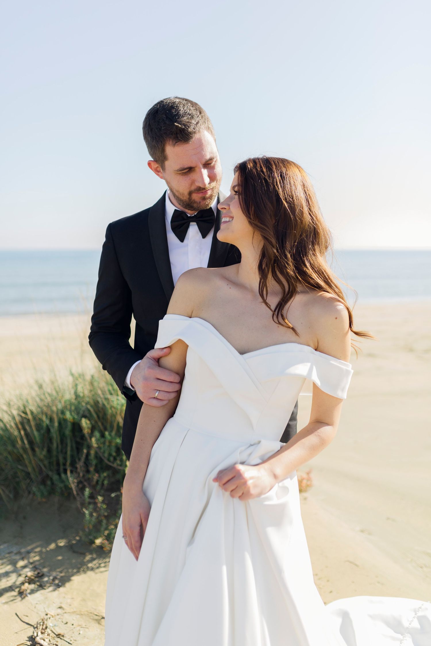 A bride and groom share a romantic embrace on a sandy beach, she in an off-shoulder white gown, he in a black tuxedo.