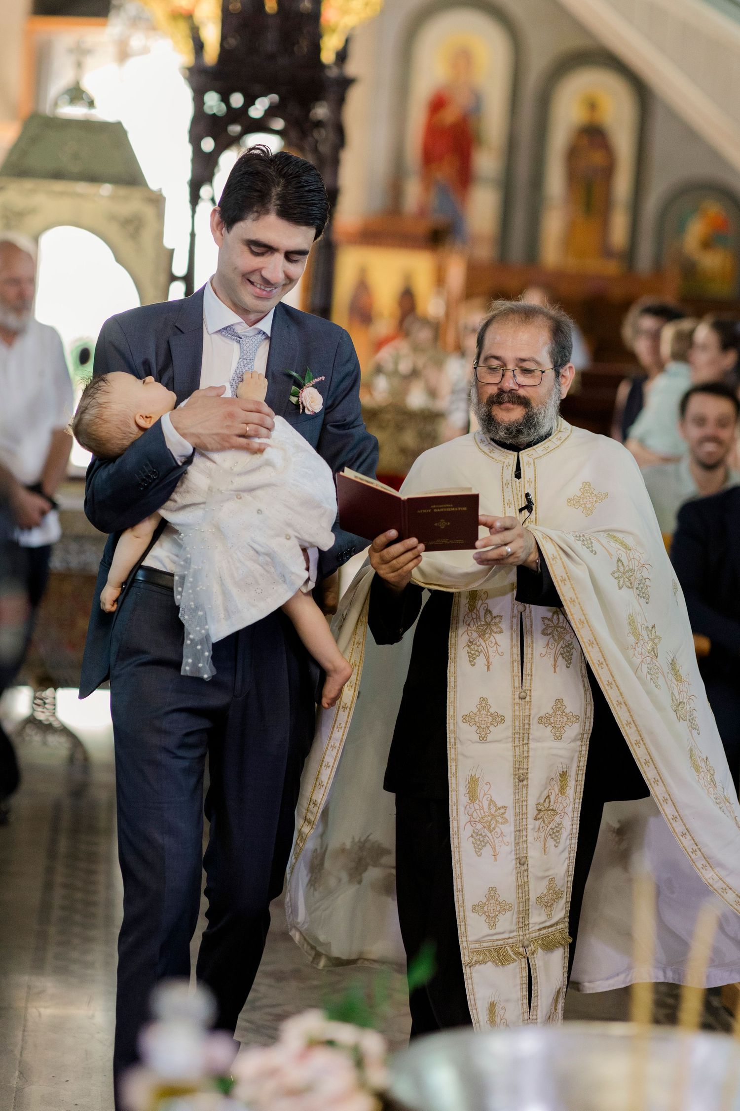 Man in suit holds baby at Orthodox baptism ceremony while priest reads from book in decorated church.