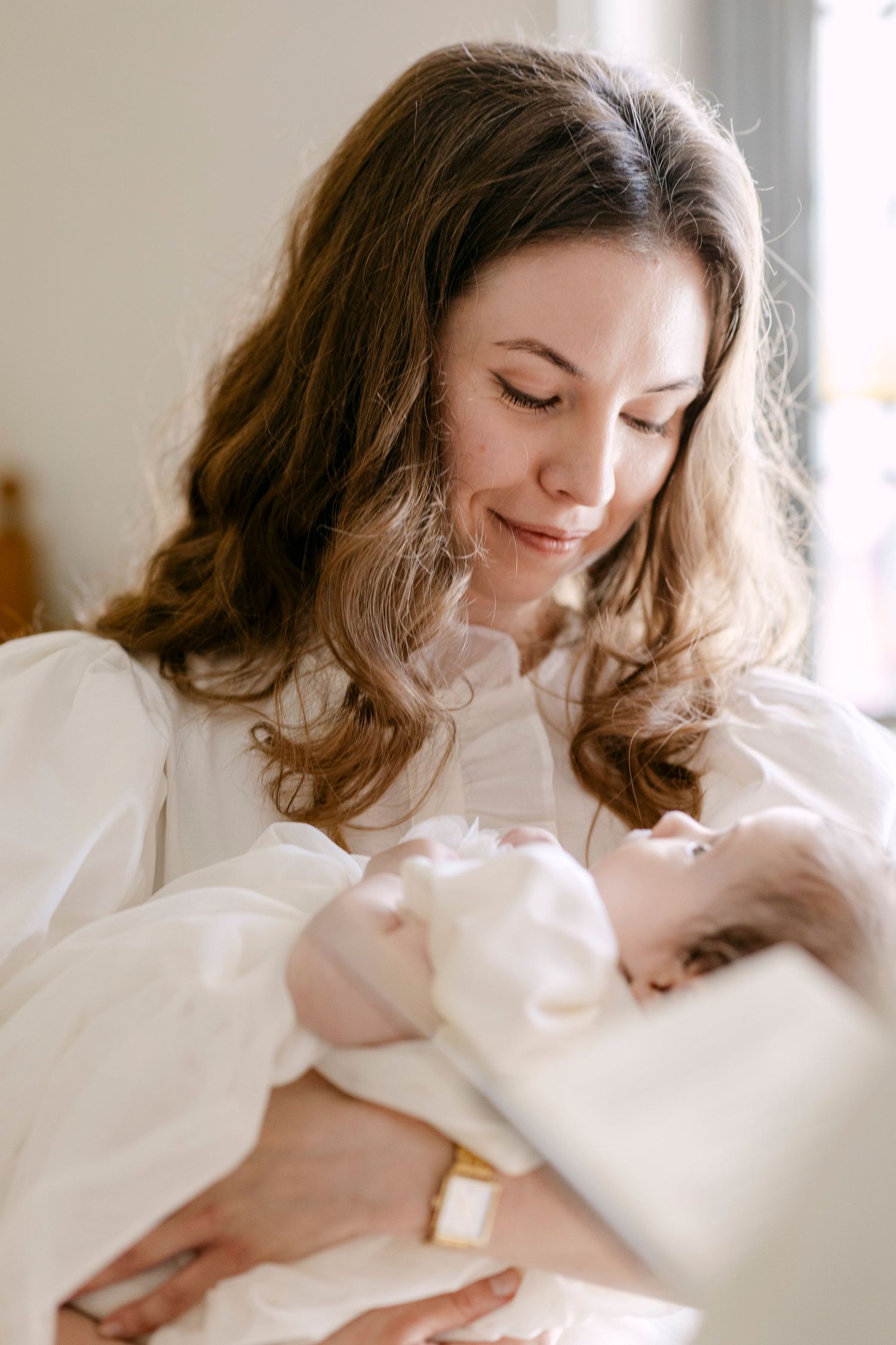 A mother with long brown hair tenderly gazes at her newborn baby wrapped in white, sharing a peaceful moment.