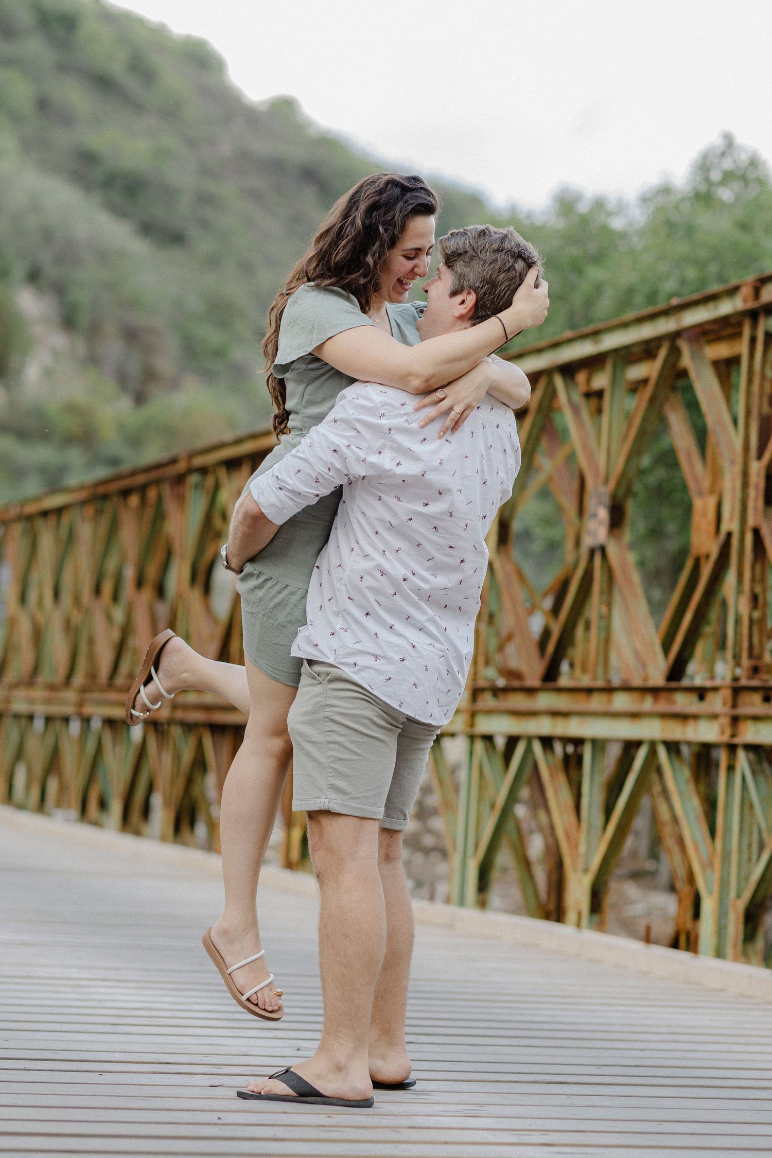 Couple embracing and smiling on a rustic bridge with green hills in the background.
