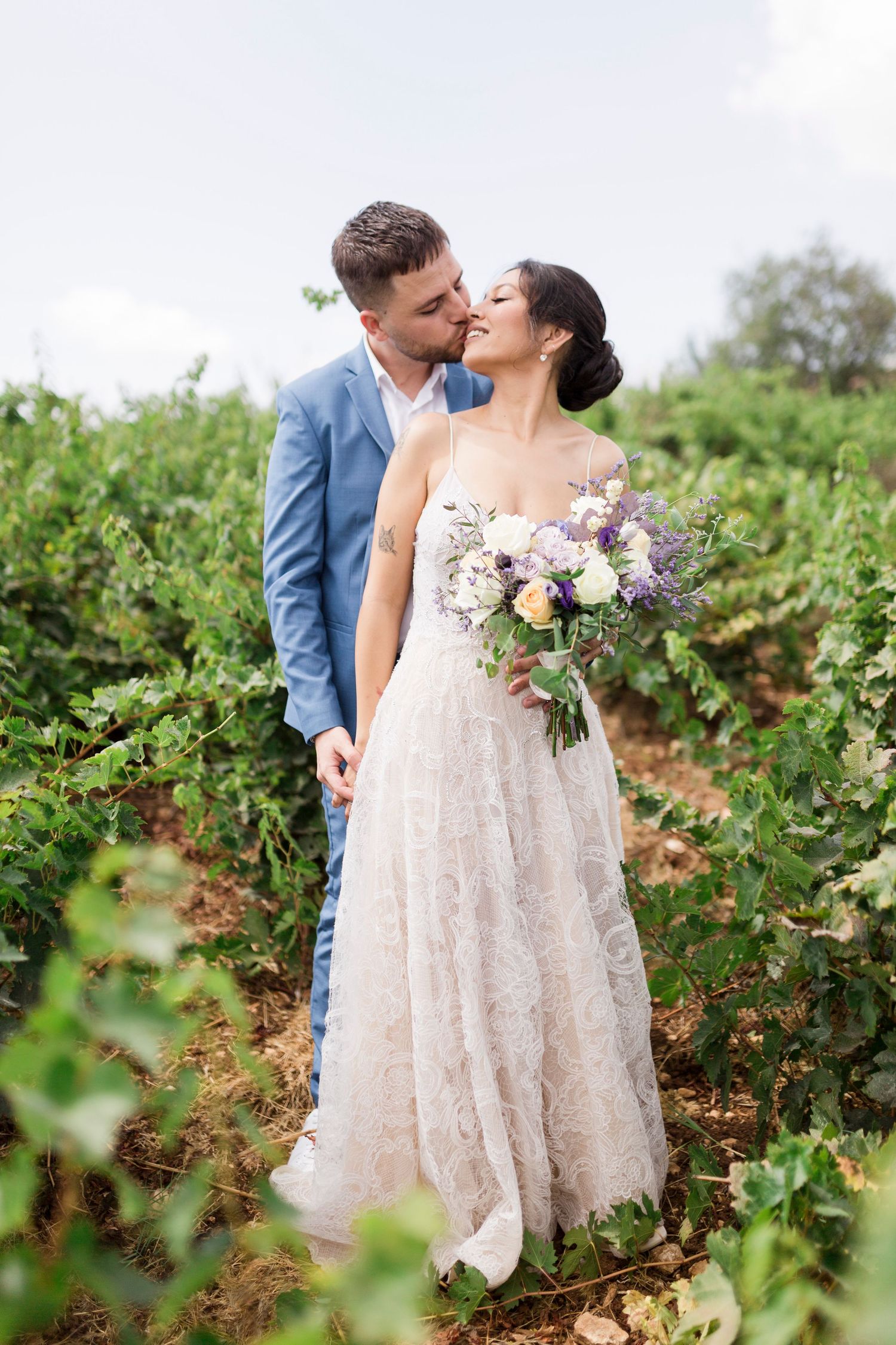 Bride and groom share a romantic kiss among green vineyard rows, bride holding purple and white bouquet.