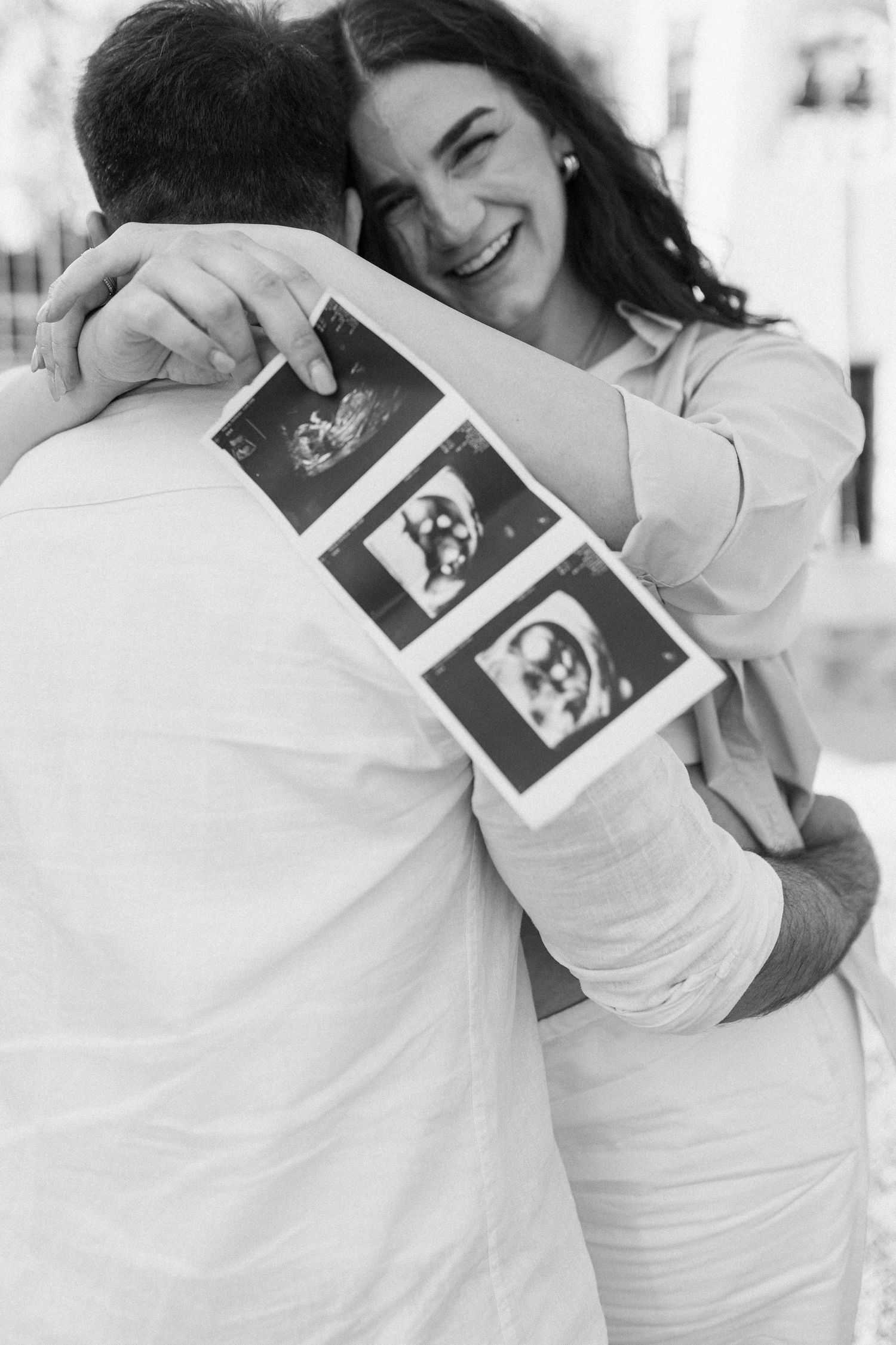Black and white photo of a joyful pregnant woman hugging her partner while holding ultrasound photos behind his back.