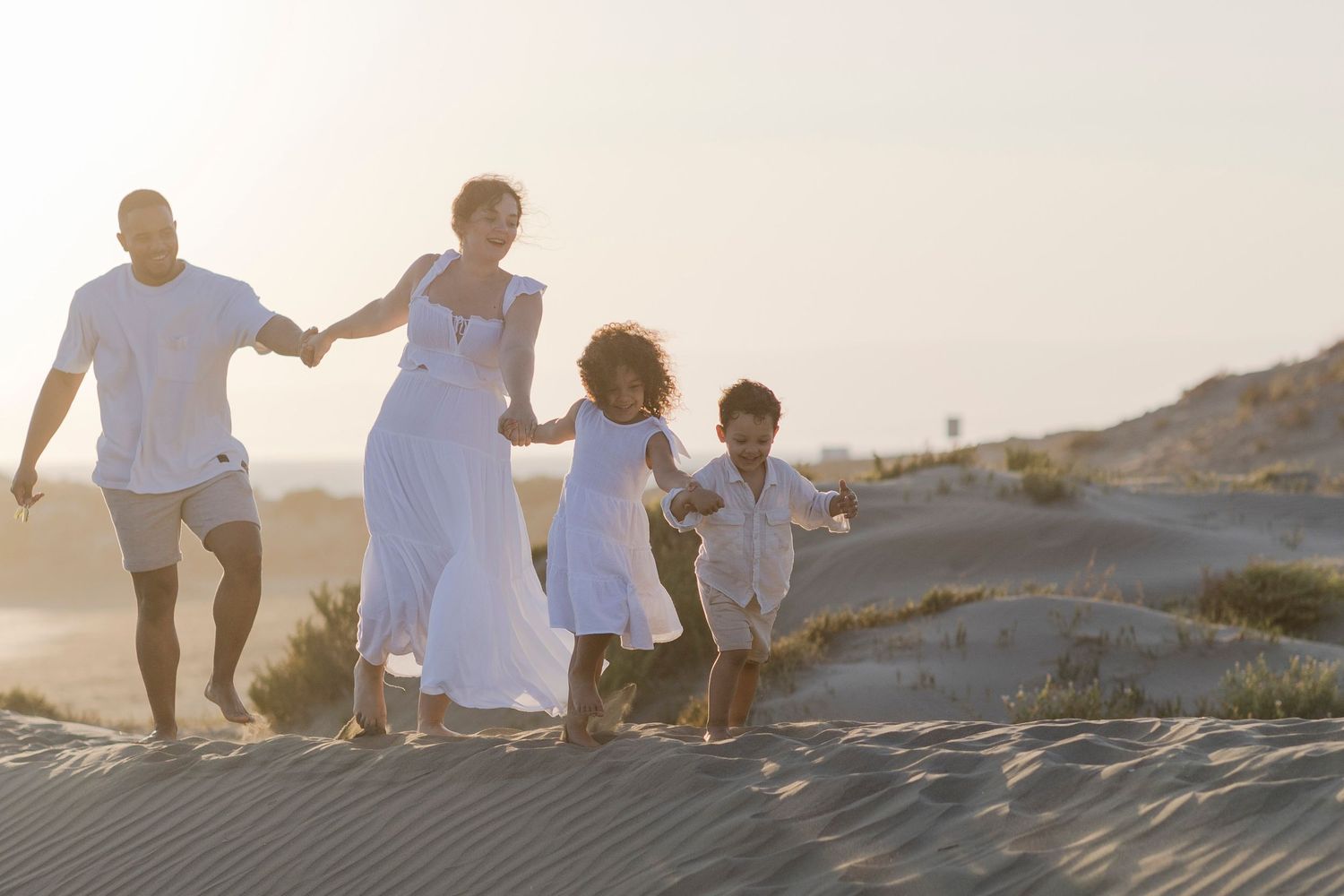 Family in white outfits running joyfully on sandy dunes at sunset.