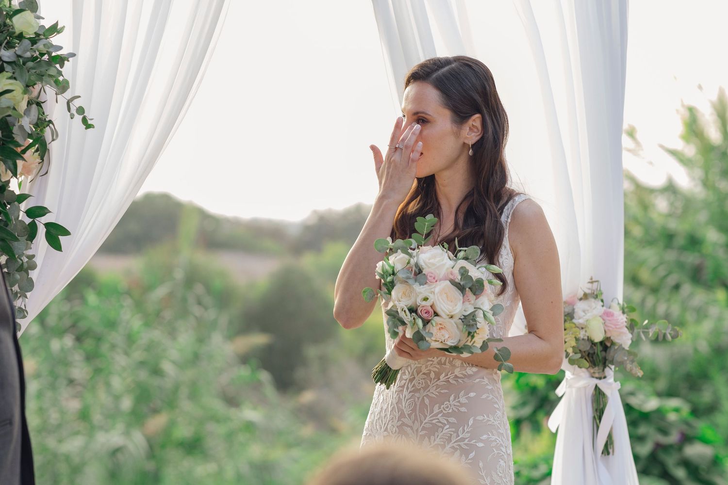 Emotional bride in sequined gown holds pink bouquet, wiping tears at outdoor wedding ceremony with white drape backdrop.