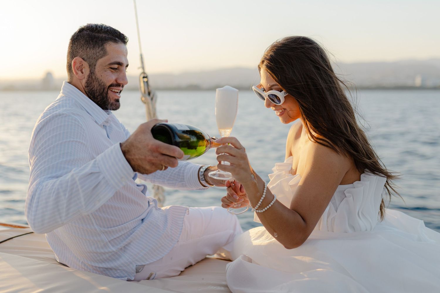 Couple in white outfits sharing champagne on a yacht at sunset, celebrating a romantic occasion.