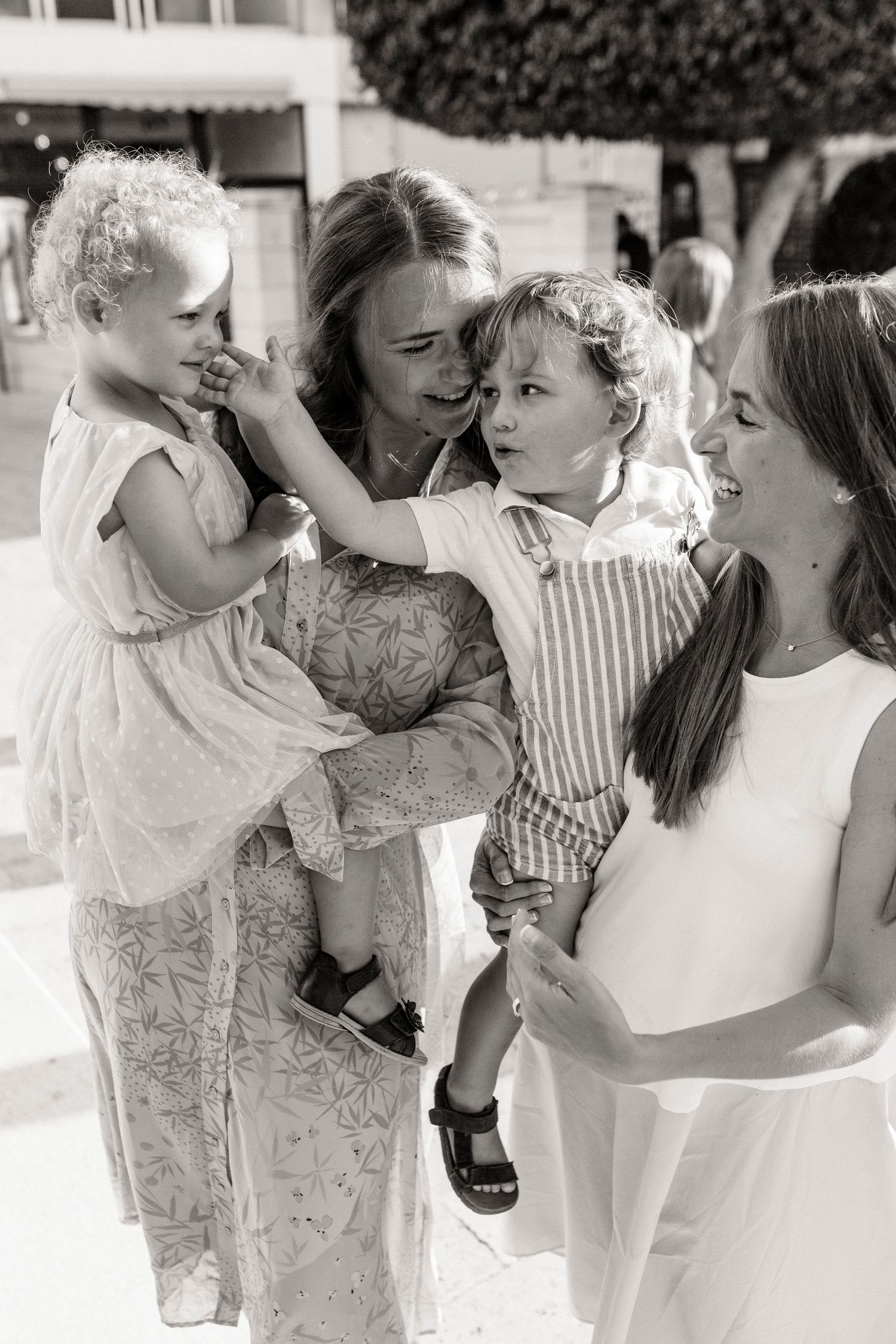 Black and white photo of two women holding toddlers outdoors, smiling and interacting at a sunny gathering.
