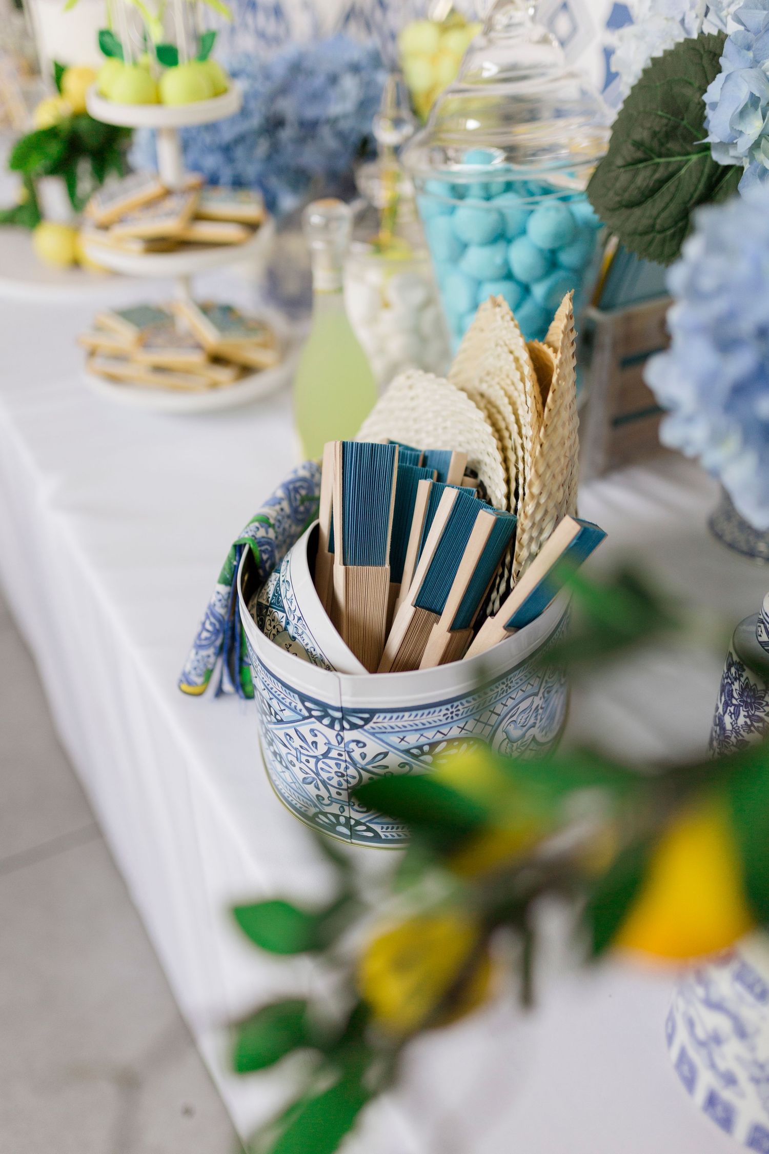 Blue and white ceramic vase holding decorative hand fans on an elegant event table with hydrangeas and candy jars.