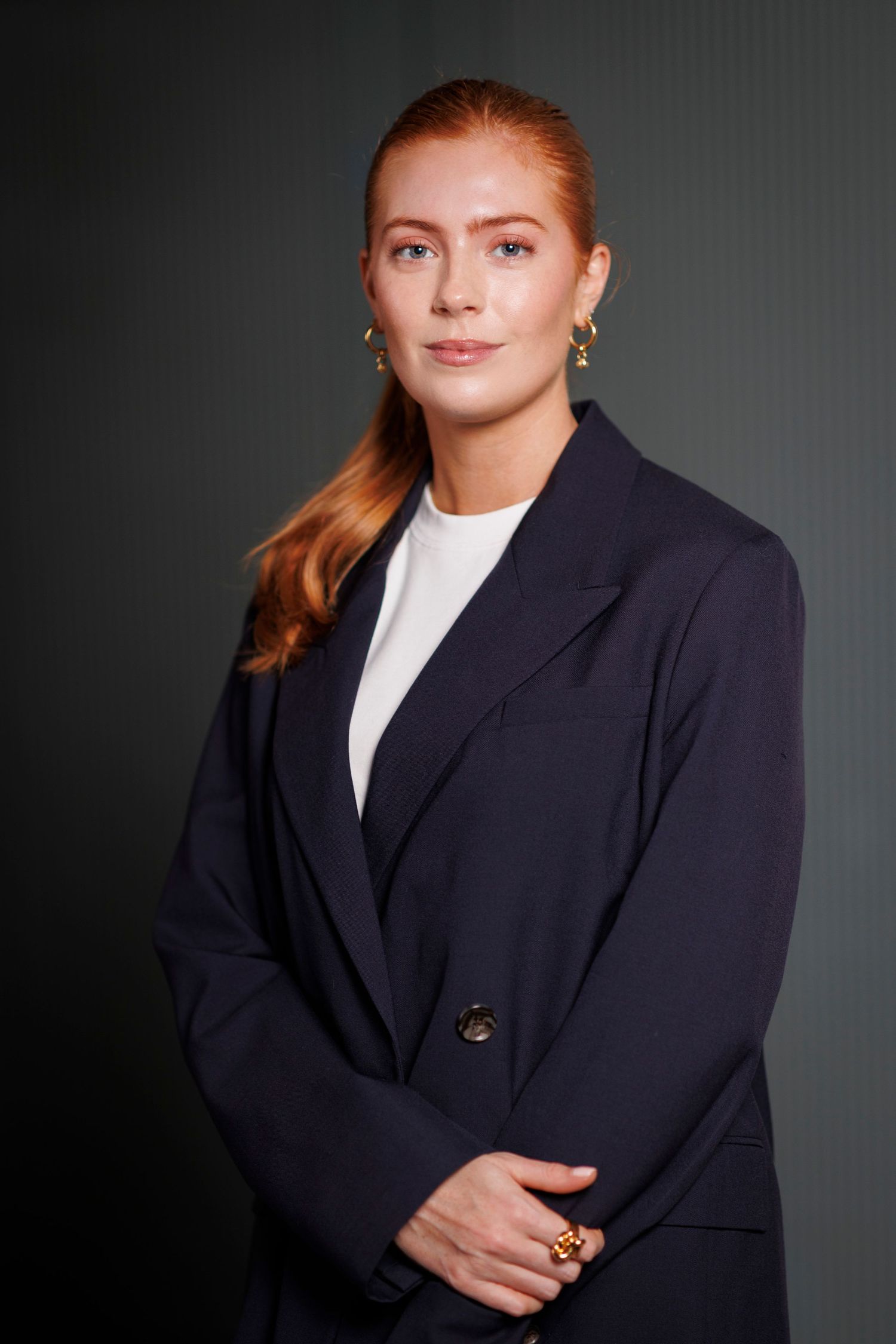 Professional woman with red hair wearing a navy blazer and gold earrings poses confidently against a dark background.