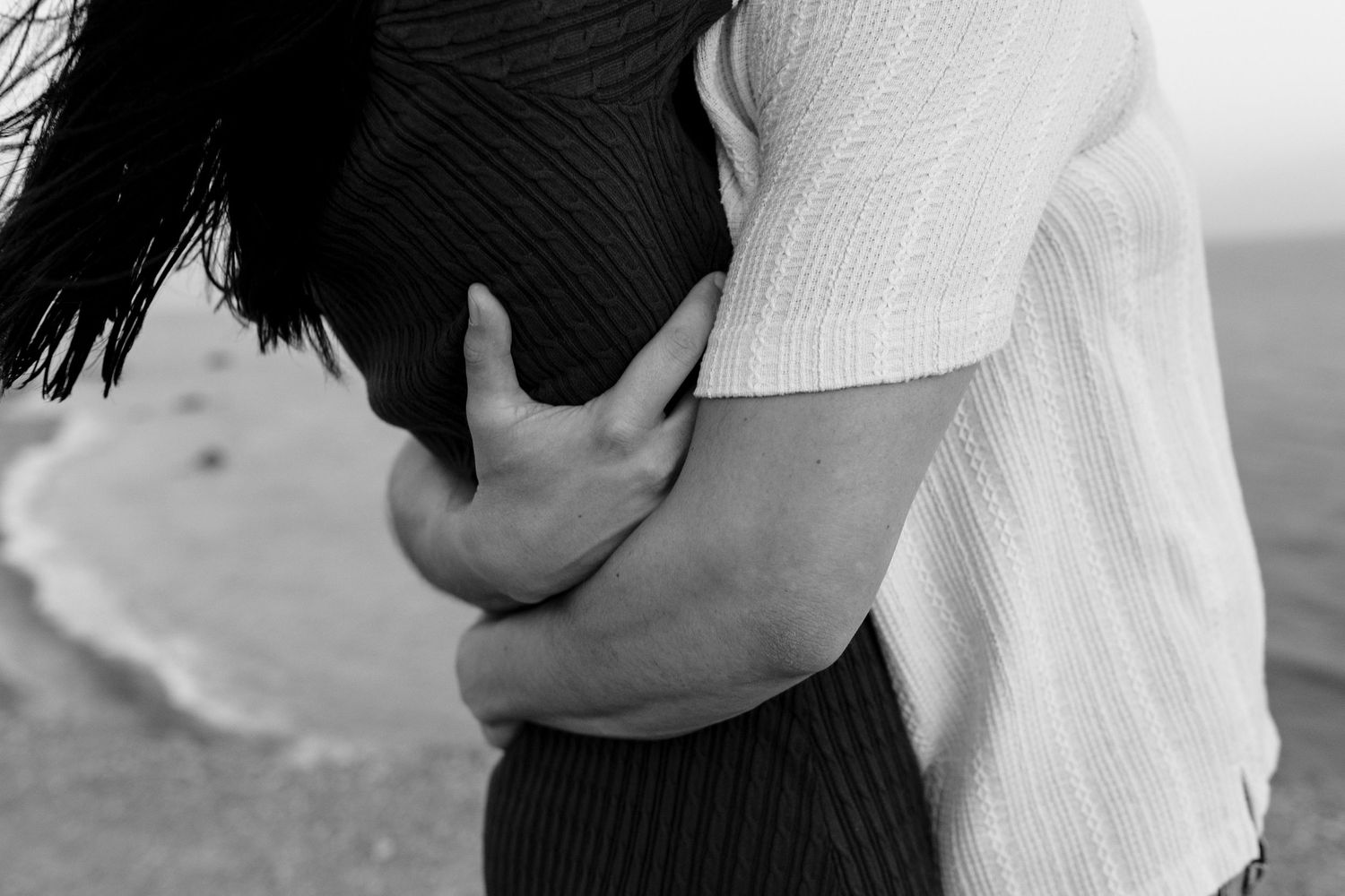 Black and white photo of a couple embracing on a beach, hands wrapped around each other tenderly.