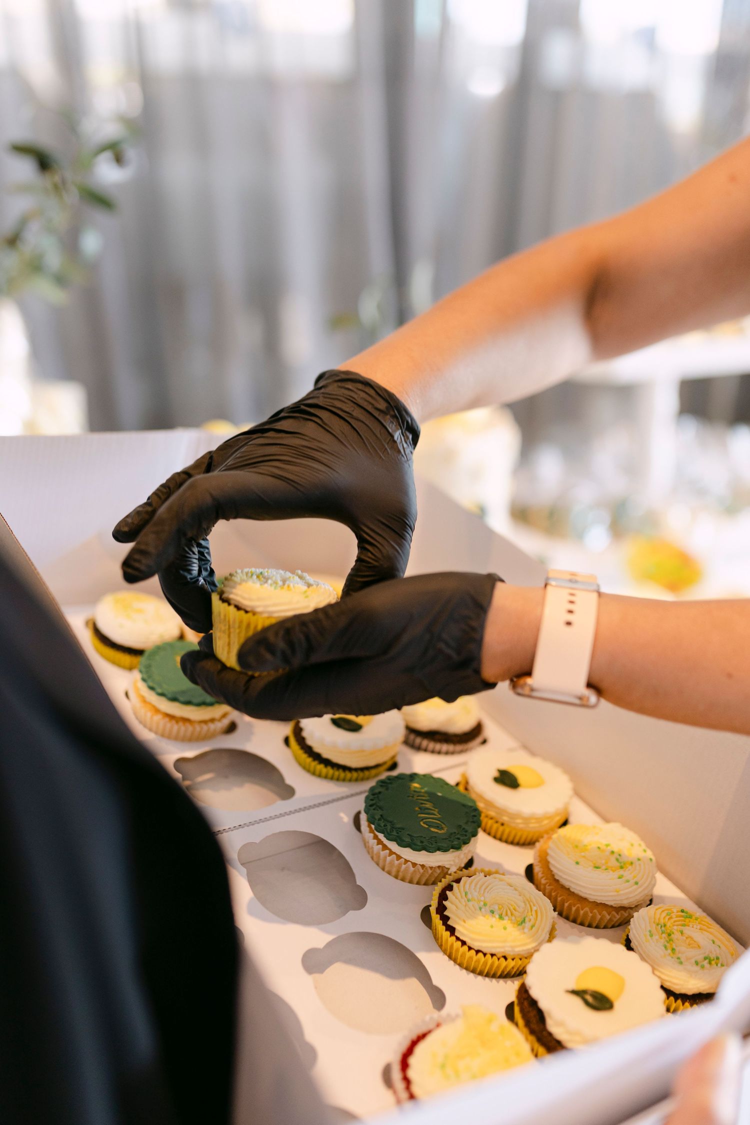 Hands in black gloves arranging colorful cupcakes in a white box at an event.