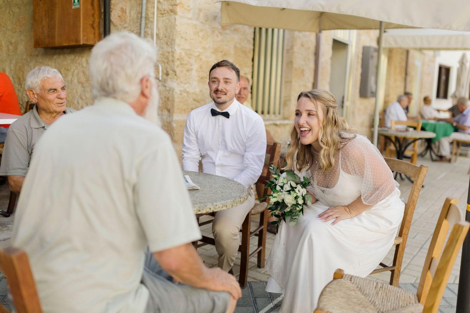 Bride and groom in wedding attire sit at outdoor cafe table, smiling and chatting with elderly guest holding bouquet.