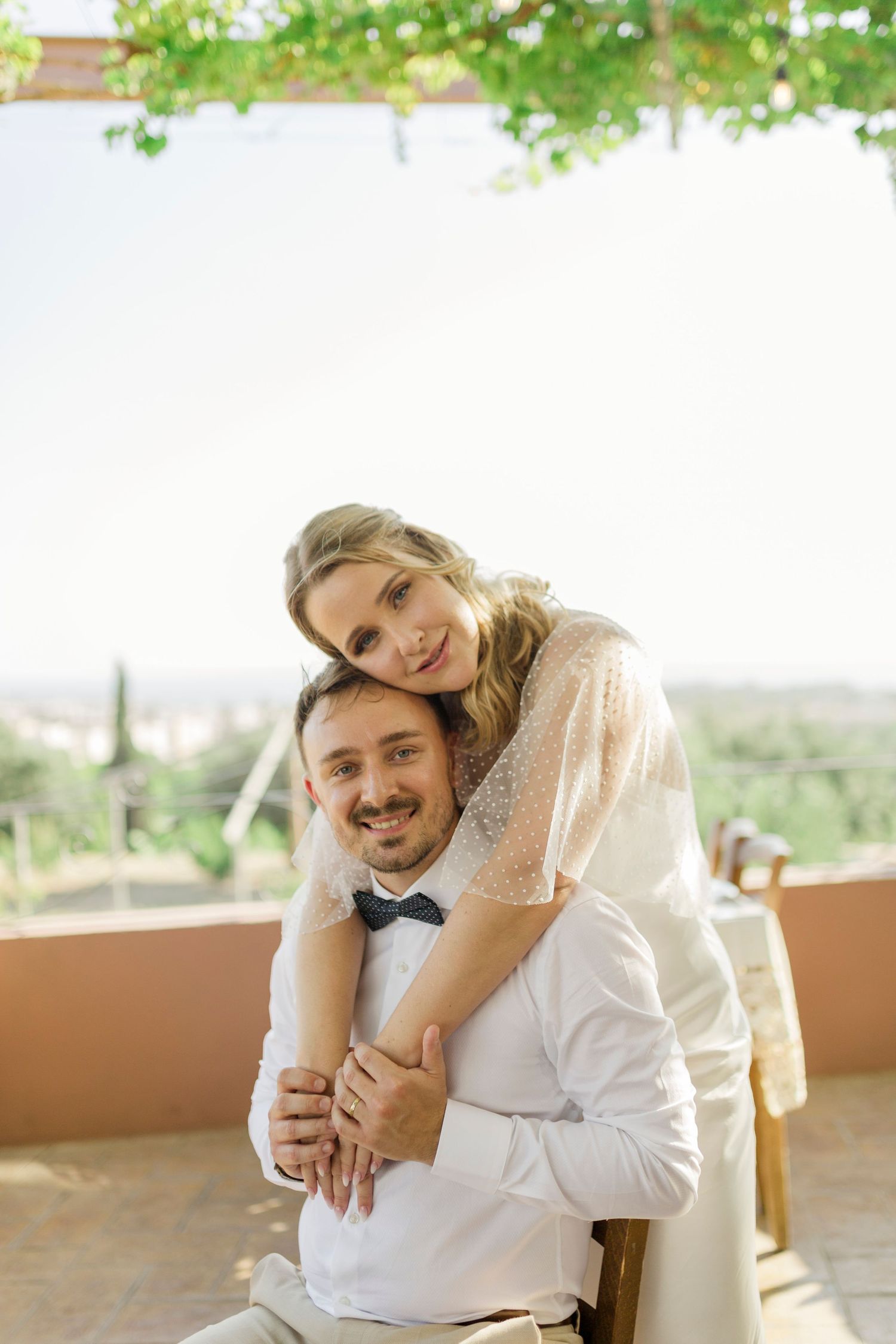 Smiling bride hugs groom from behind at outdoor wedding venue with scenic views.