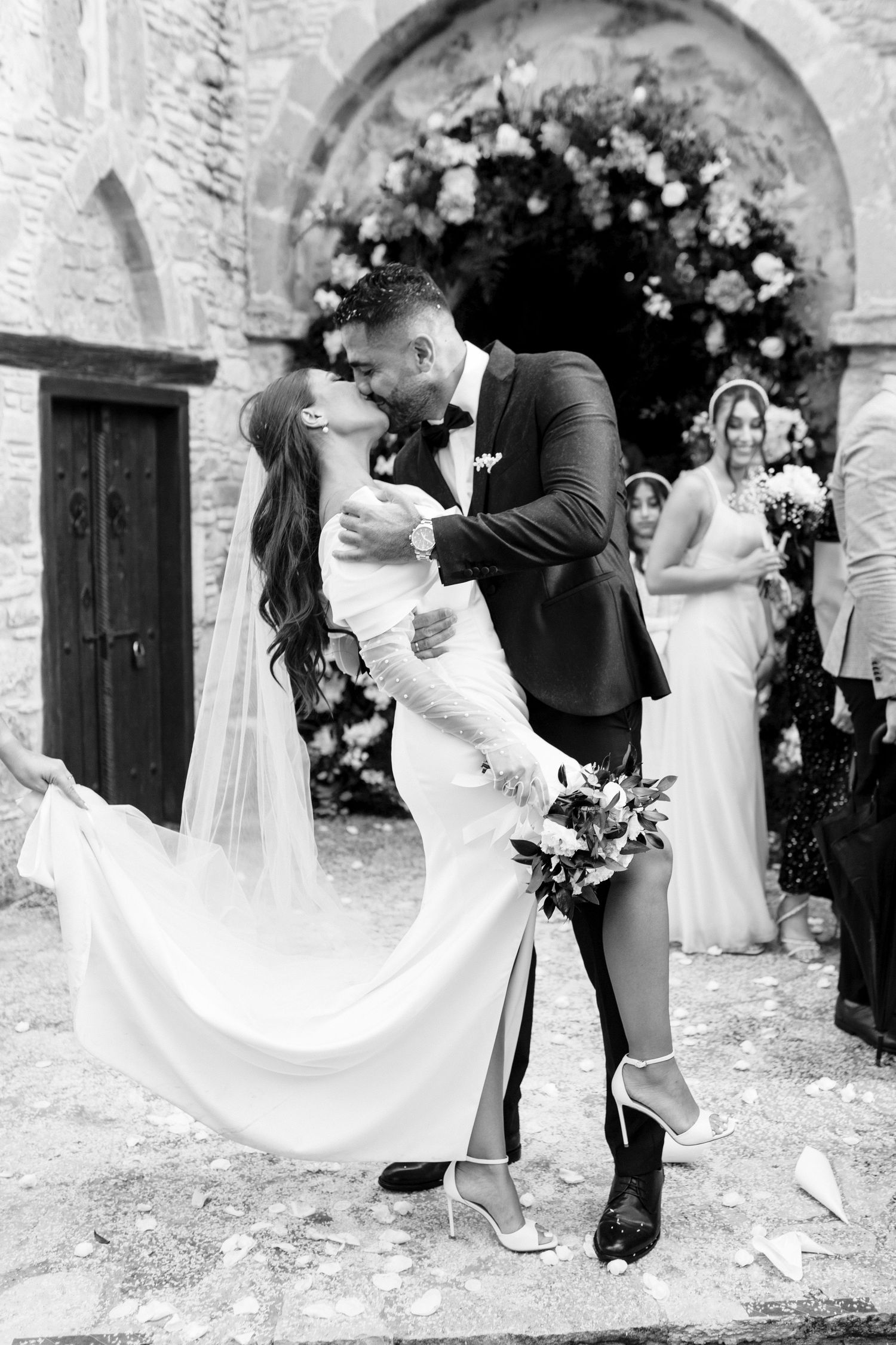 Black and white photo of bride and groom sharing a romantic kiss outside stone church after wedding ceremony.