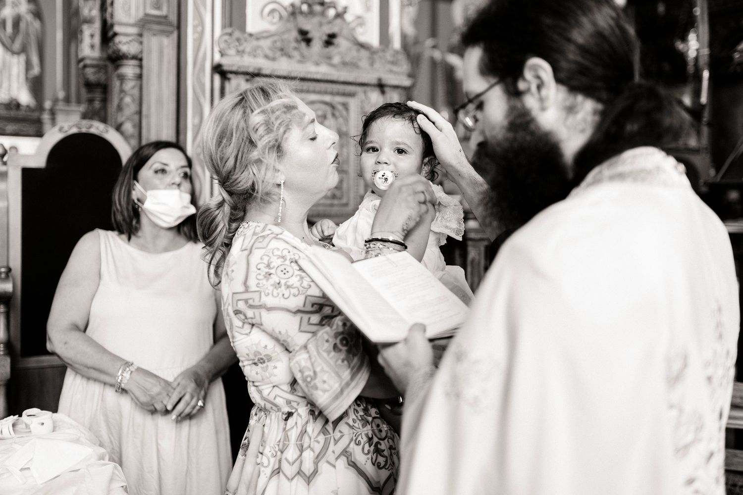 Black and white photo of a baby's baptism ceremony with a priest, godmother, and family in a church.