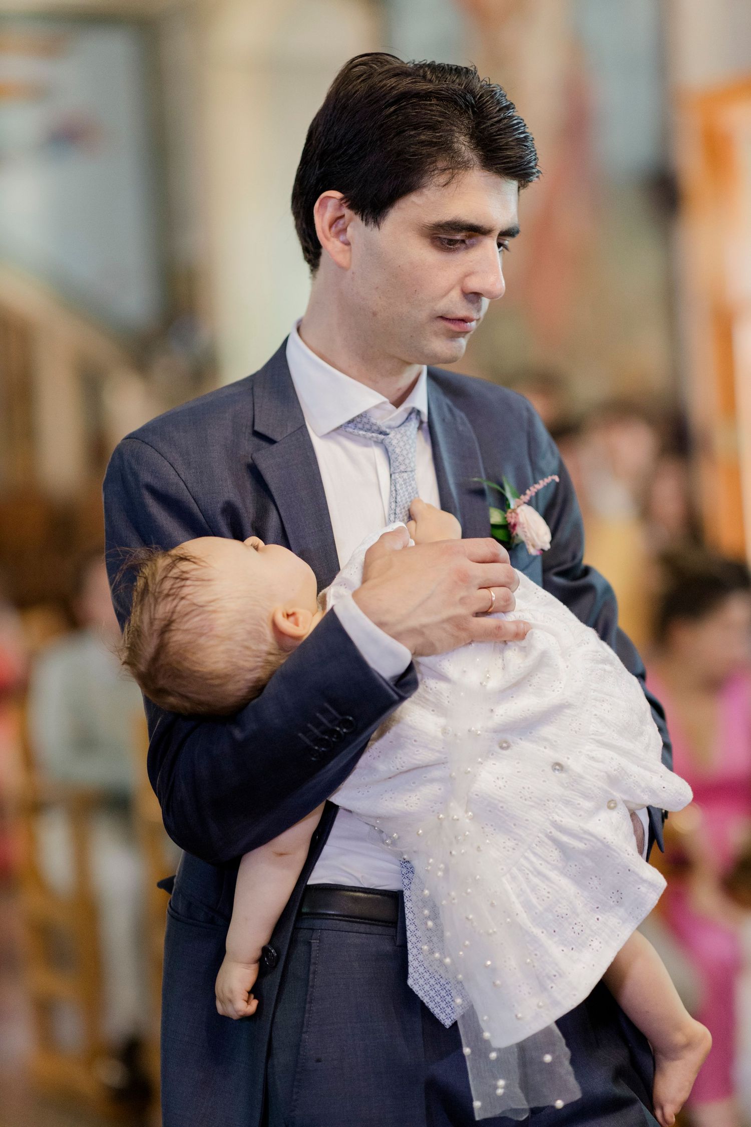 Man in suit holding a baby dressed in white christening gown during a baptism ceremony.