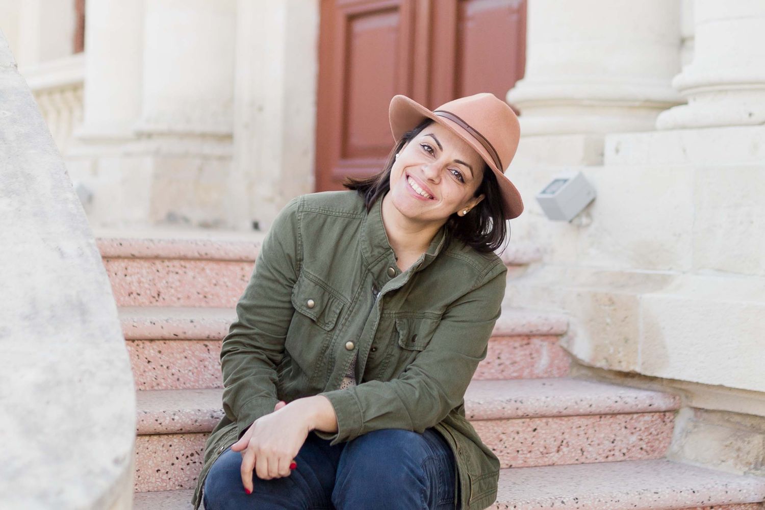 Smiling woman in green jacket and brown hat sitting on stone steps outdoors.