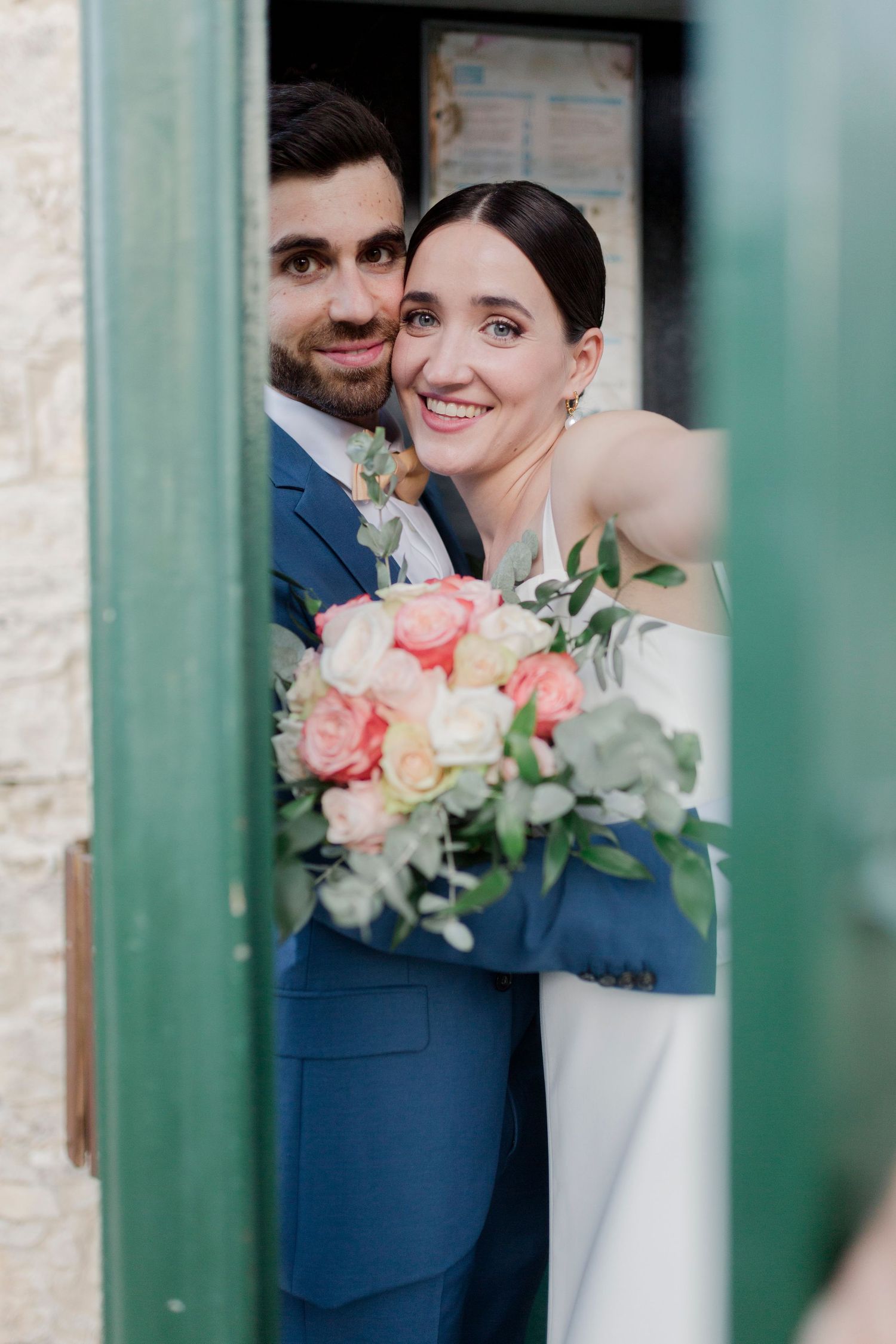 Smiling bride holding pink and white rose bouquet with groom in blue suit peeking through green doorway.