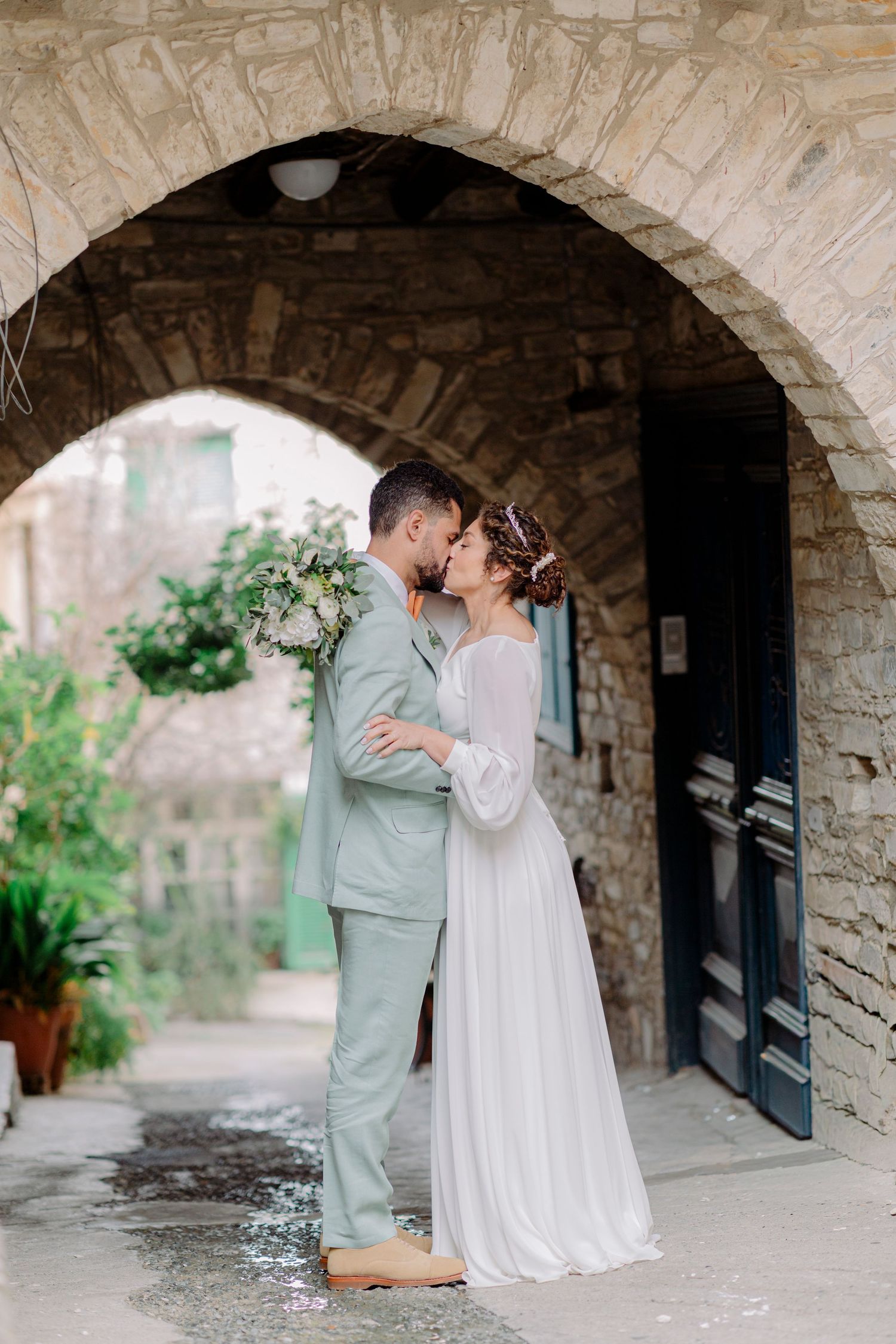 Bride and groom share a romantic kiss under a stone arch at a rustic European venue.
