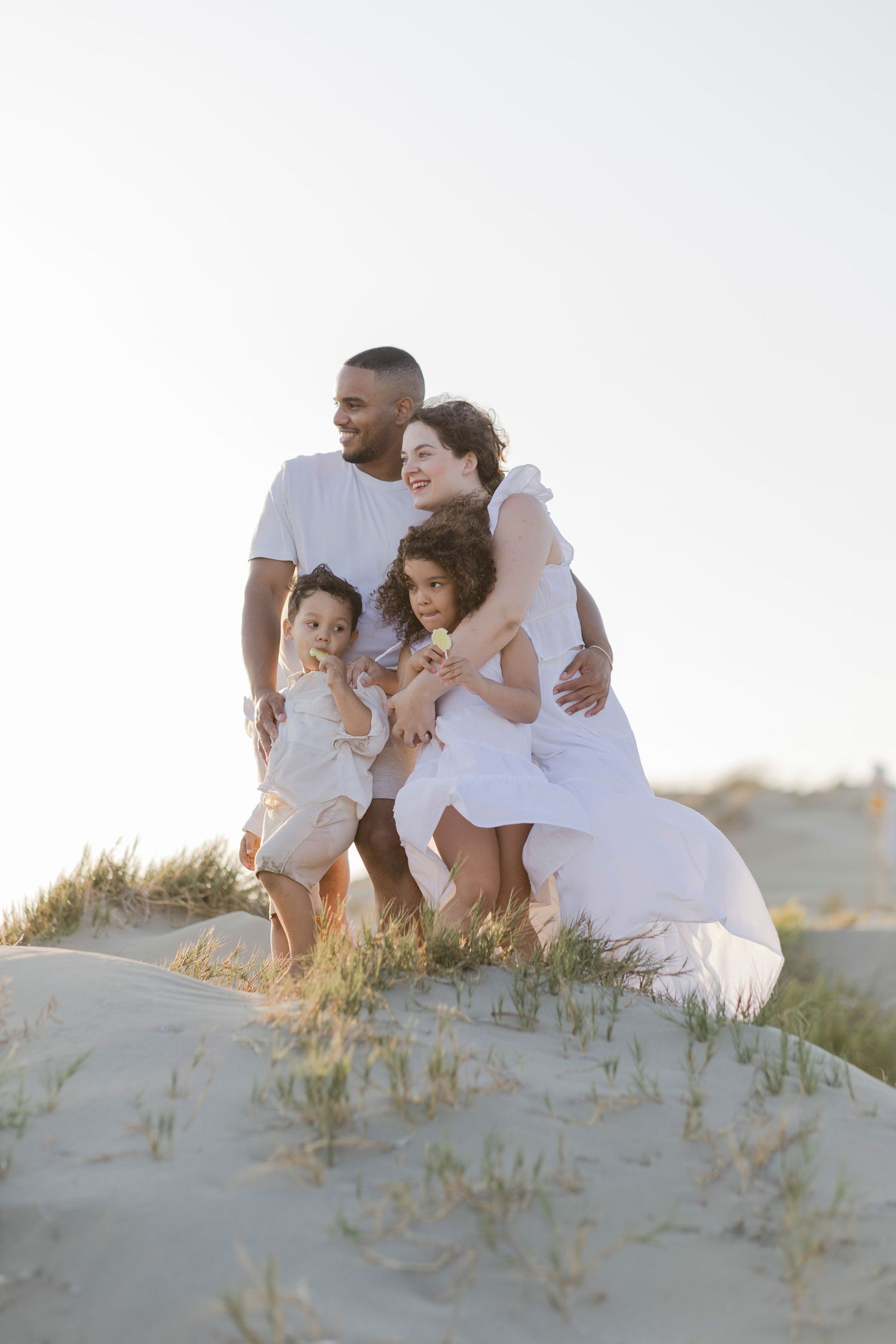 Happy family of four in white outfits posing together on sandy dunes during golden hour at the beach.