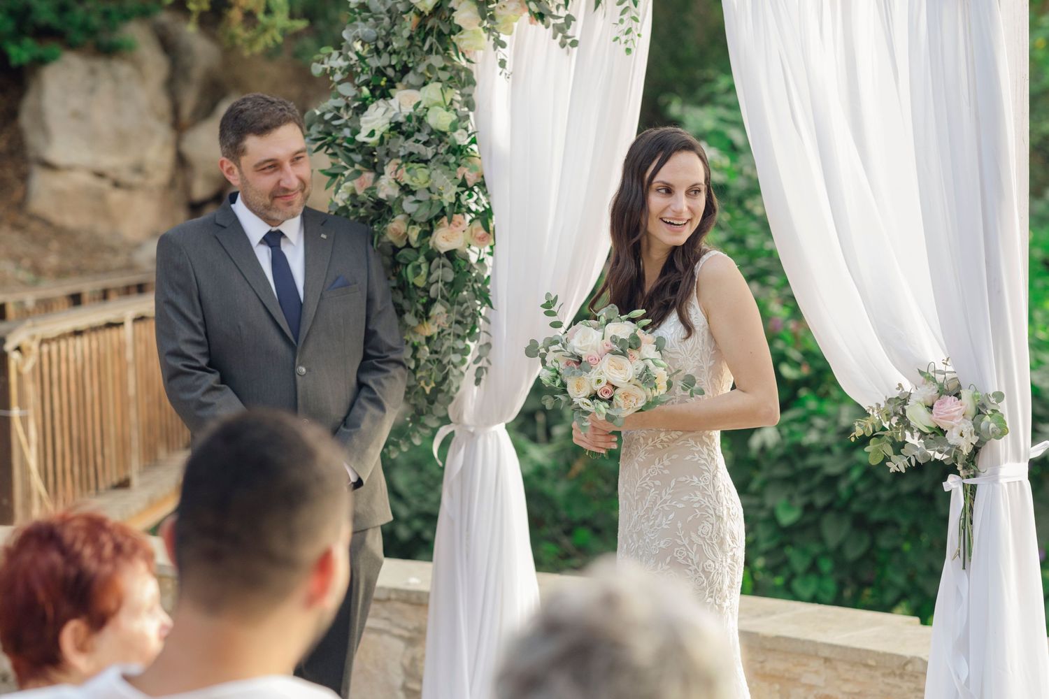 Bride holding bouquet smiles at groom during outdoor wedding ceremony with white draping and floral arch.