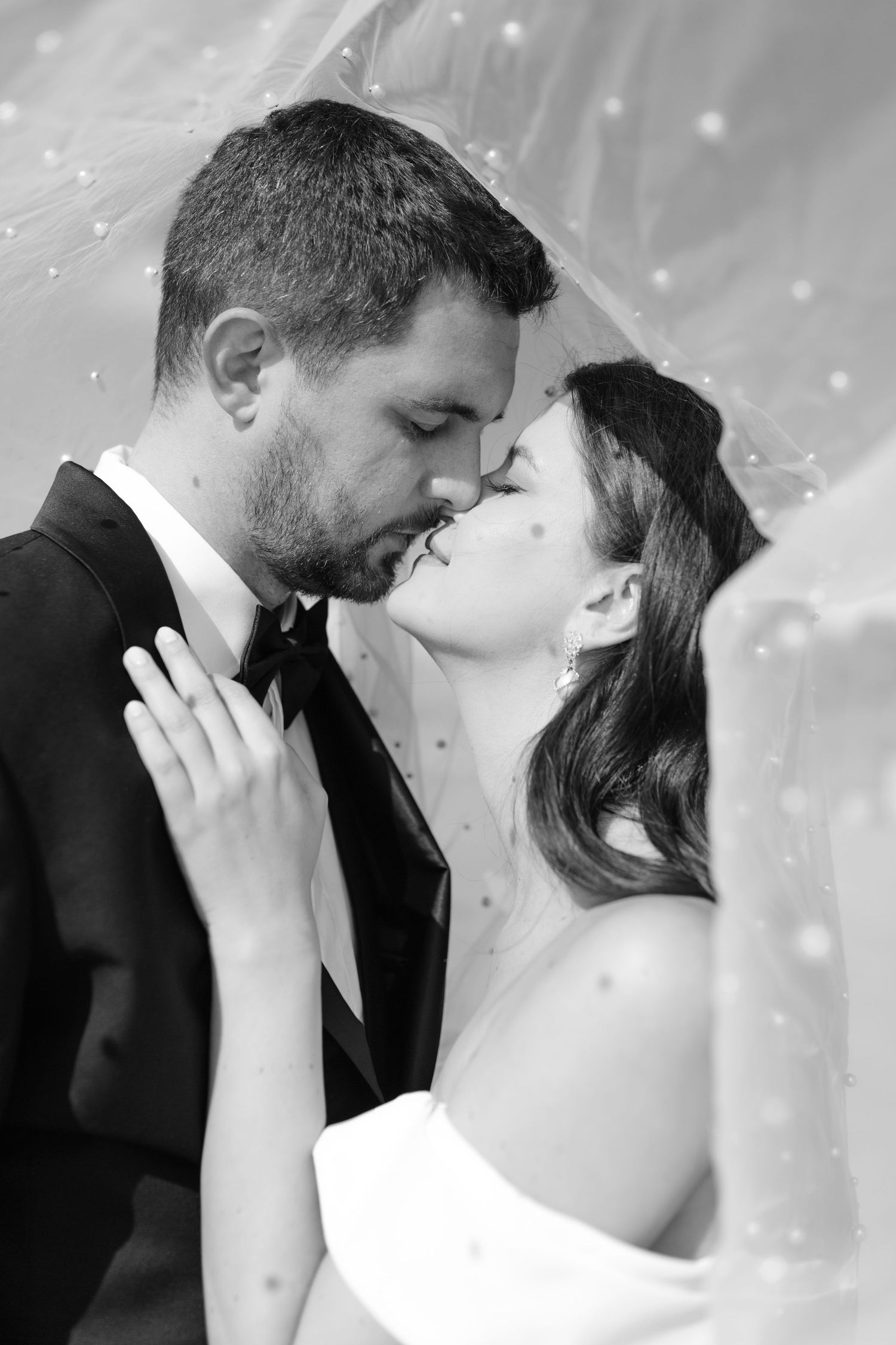 Black and white photo of bride and groom sharing a tender kiss under a pearl-adorned wedding veil.