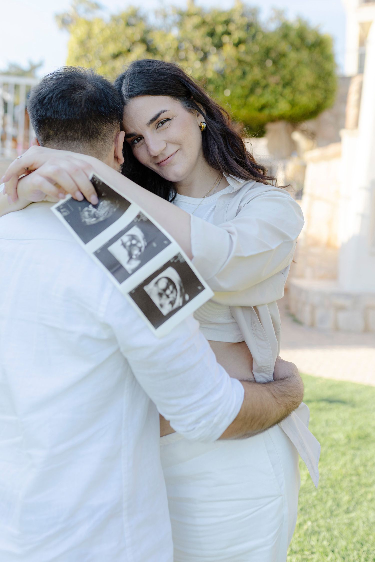 Pregnant woman in white dress hugs man outdoors, holding ultrasound photos, smiling warmly in a garden setting.