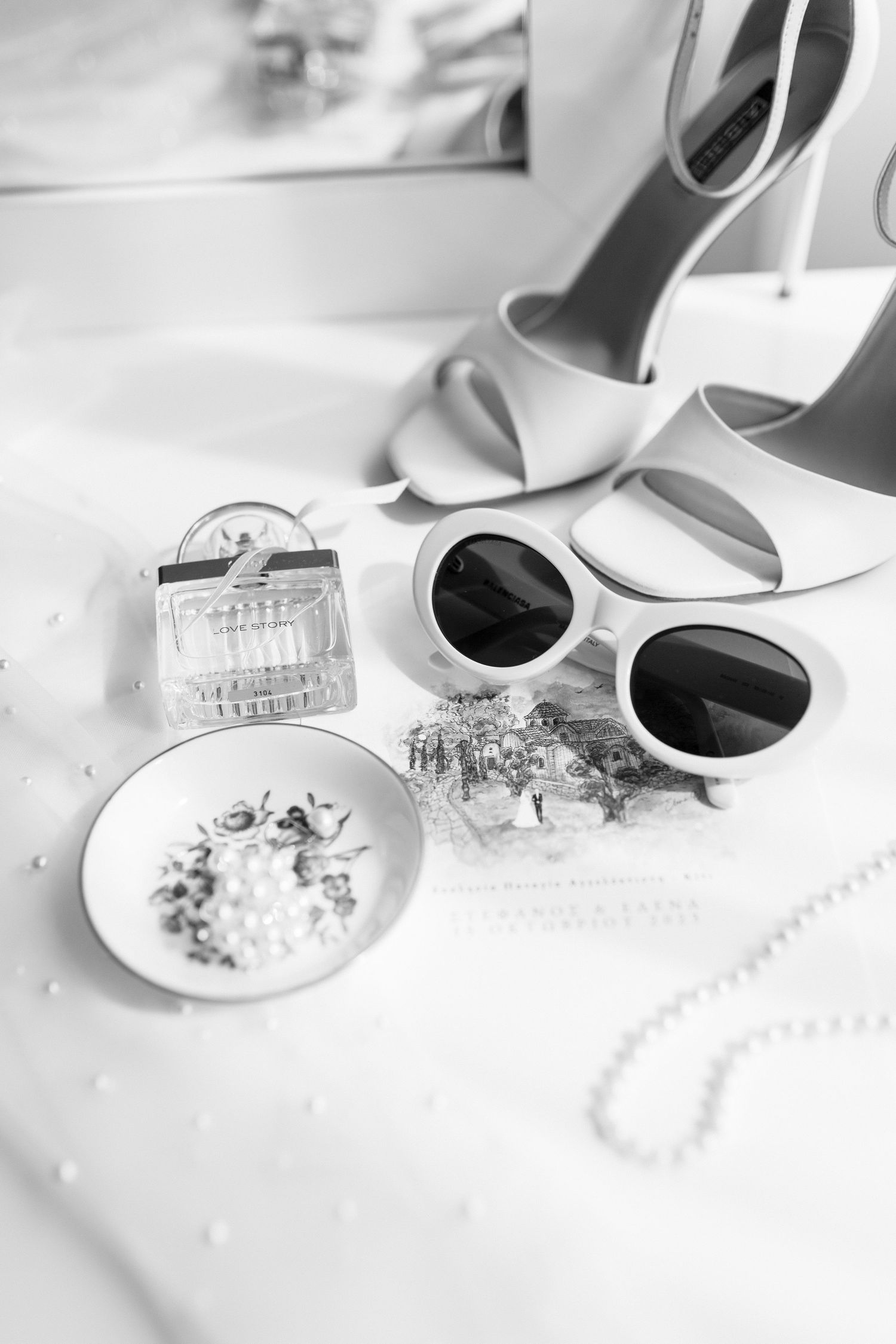 Elegant black and white bridal flat lay featuring high heels, sunglasses, perfume bottle, and pearl jewelry on white fabric.
