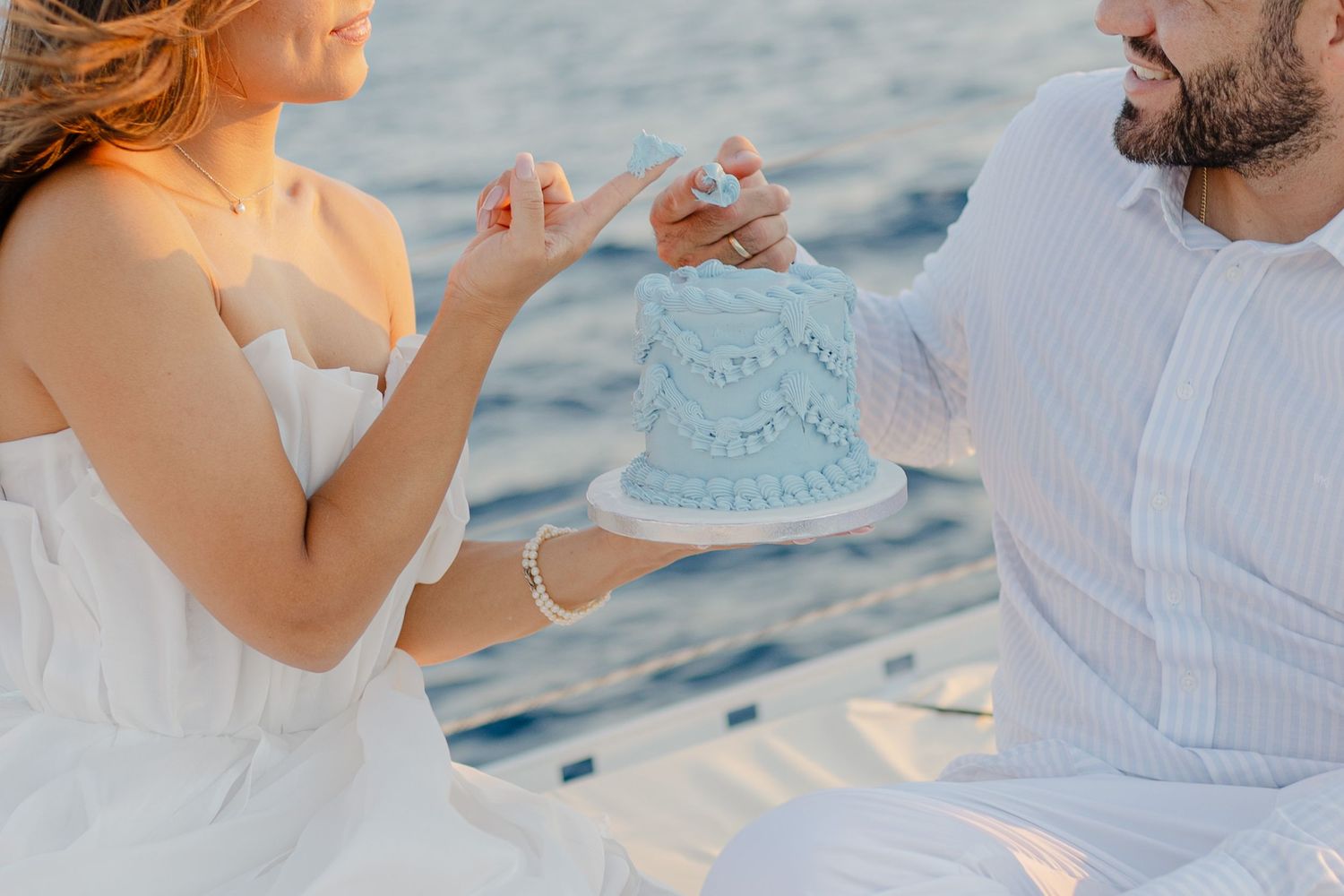 Couple sharing a small blue wedding cake outdoors near water, dressed in white for a romantic celebration.