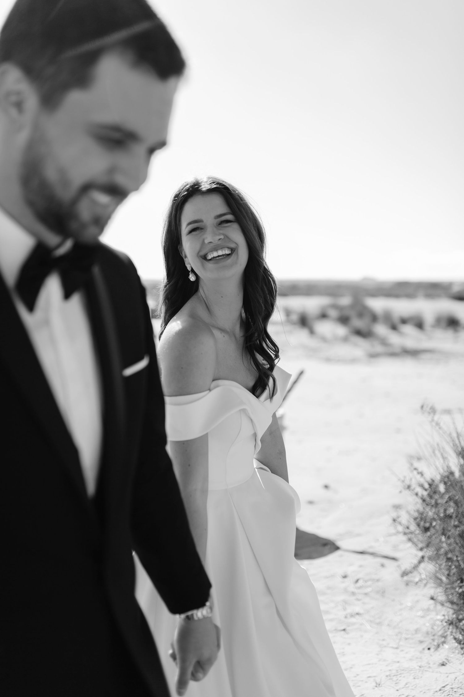 Black and white photo of a smiling bride in off-shoulder gown holding hands with groom in tuxedo on sandy beach.