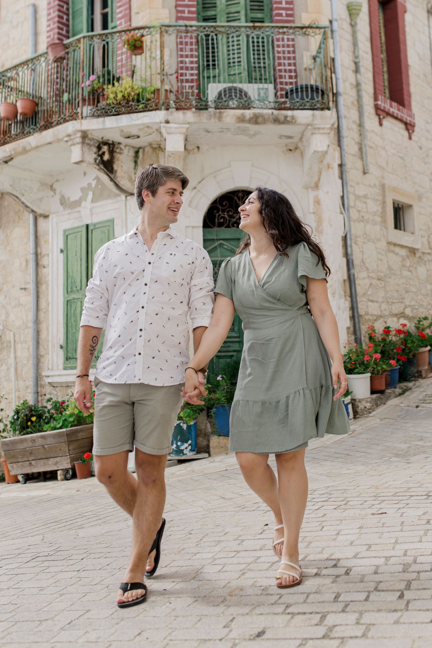 Couple holding hands walking on cobblestone street in Mediterranean town with stone buildings and green shutters.