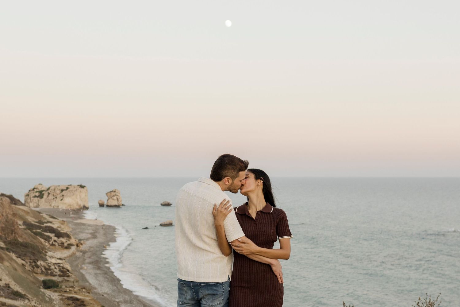 Couple sharing a romantic kiss at sunset by the sea, with rocky cliffs and a full moon in the pastel sky.