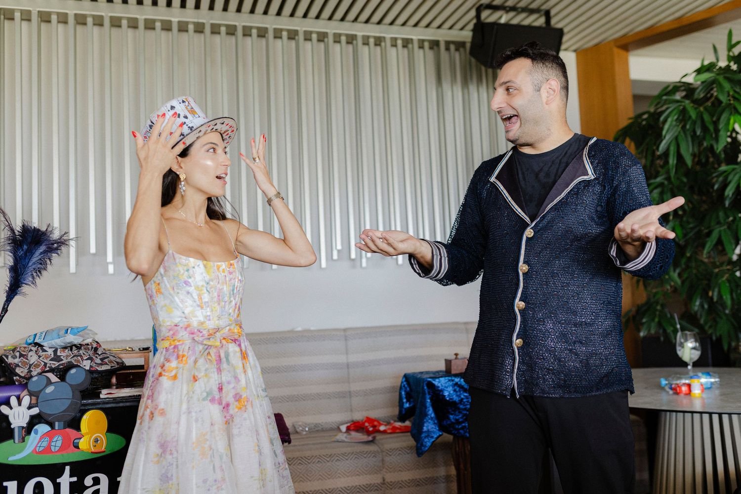 Woman in floral gown tries on hat while man in dark jacket gestures expressively at indoor party event.