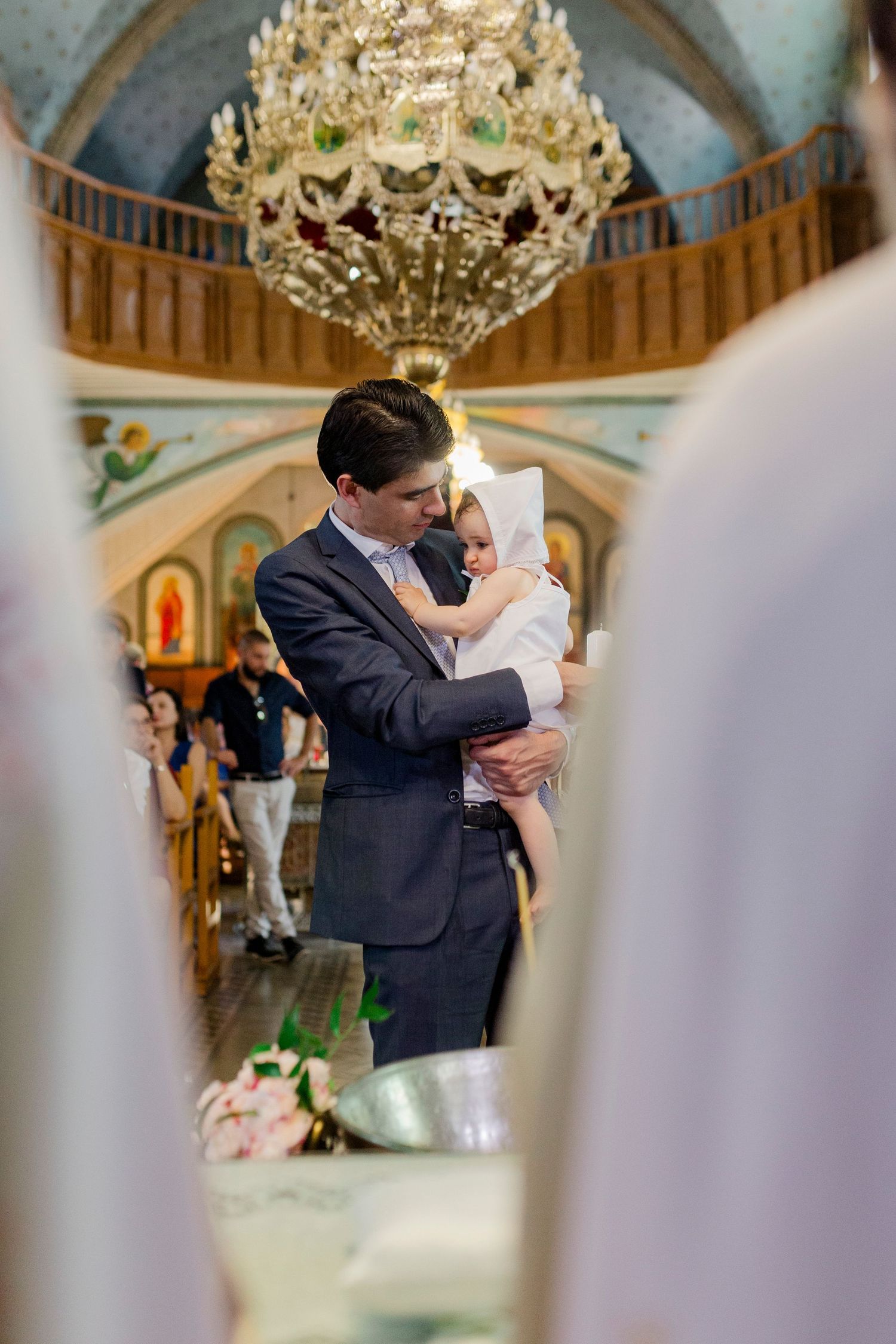 Man in suit holding baby in white christening gown inside ornate church with crystal chandelier.