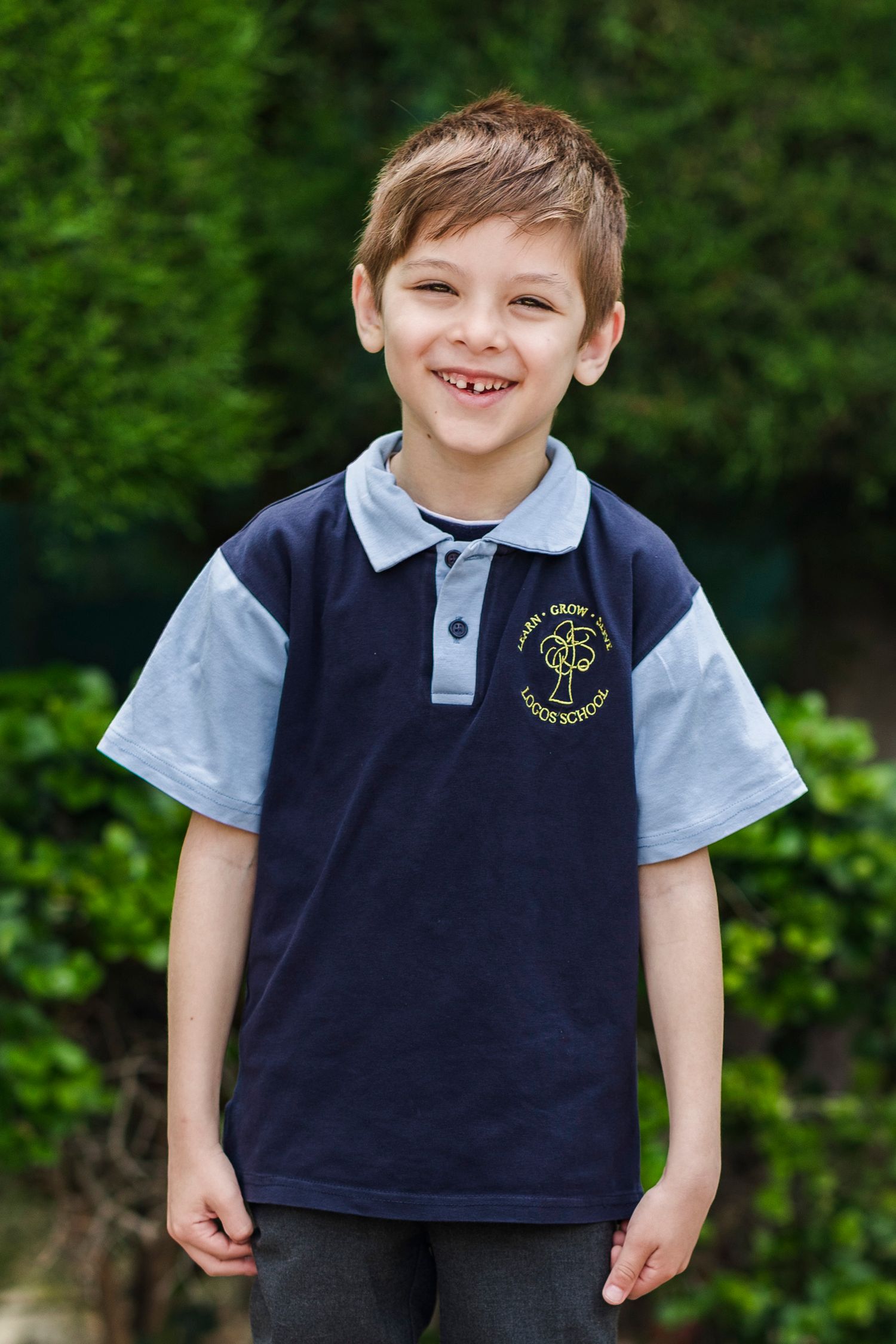 Smiling young boy wearing a navy and grey school polo shirt with emblem, standing outdoors.