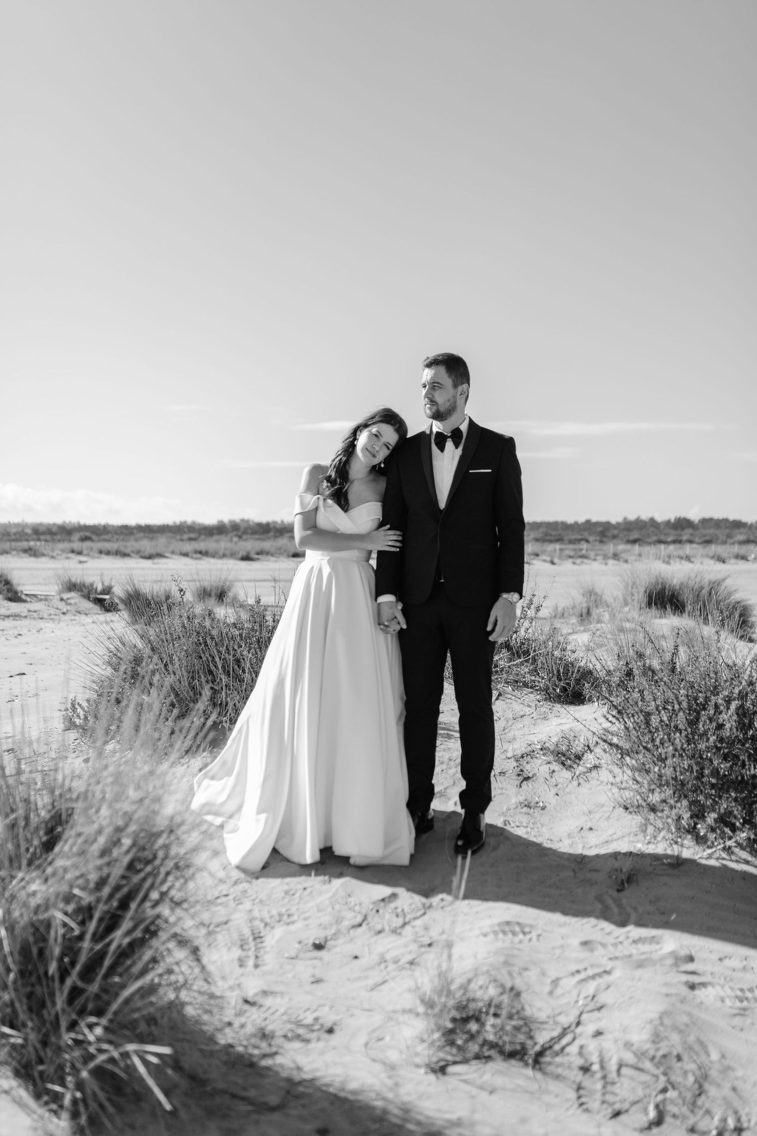 Black and white photo of bride leaning on groom in tuxedo while standing together on sandy dunes.