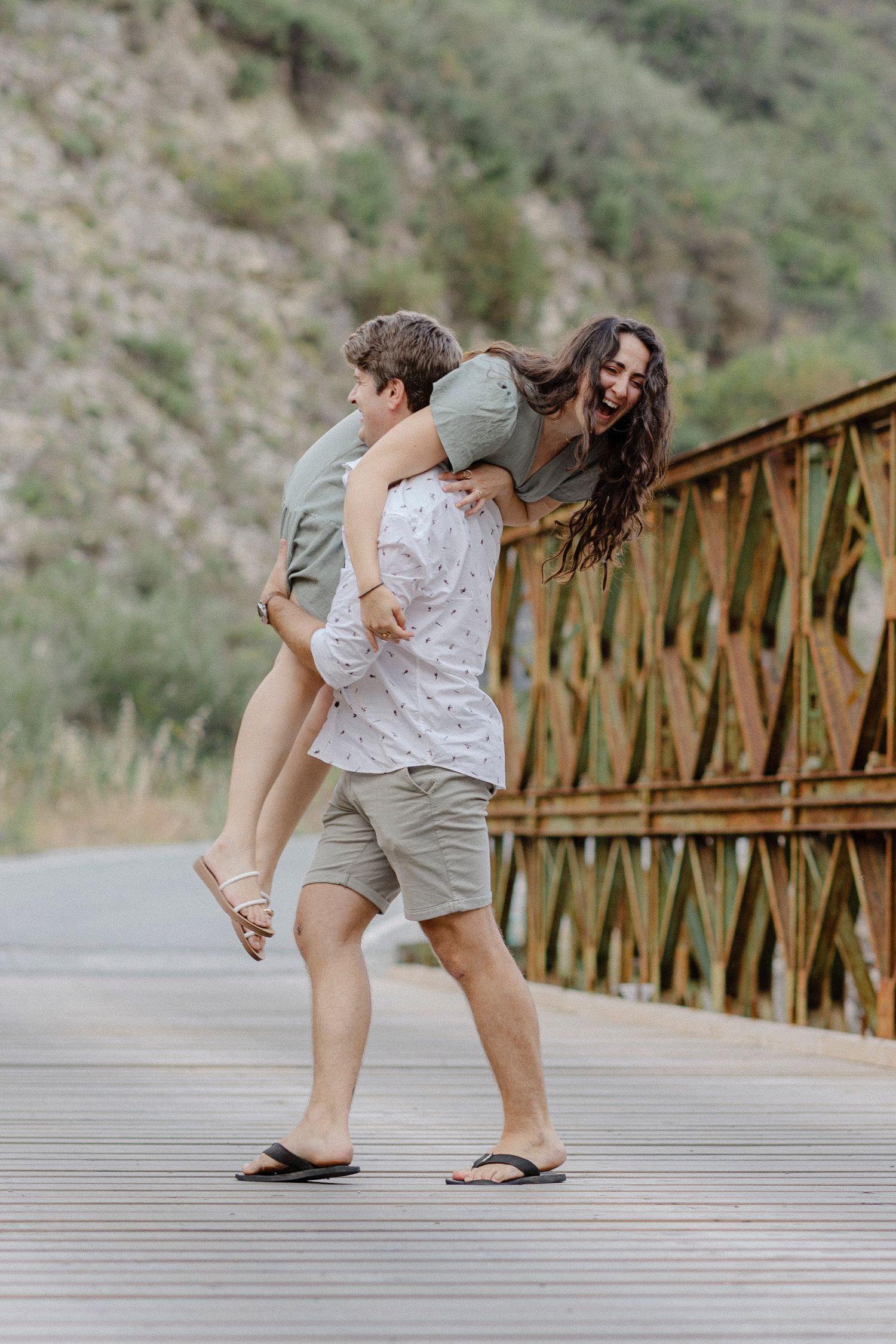 A joyful couple laughing on a bridge, man giving woman a piggyback ride with green hills in background.