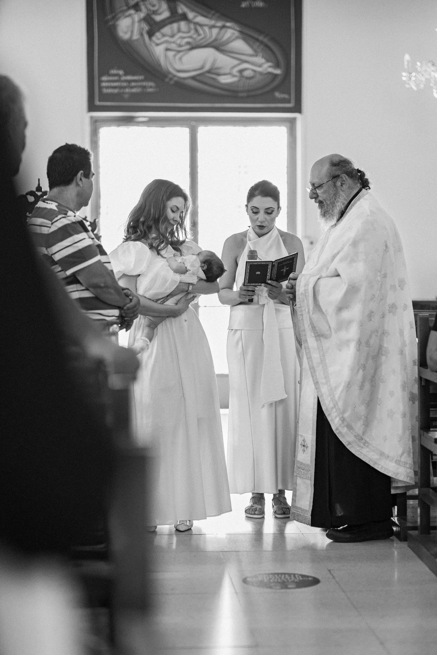 Black and white photo of a baptism ceremony with priest, godmothers in white dresses, and infant in a church.
