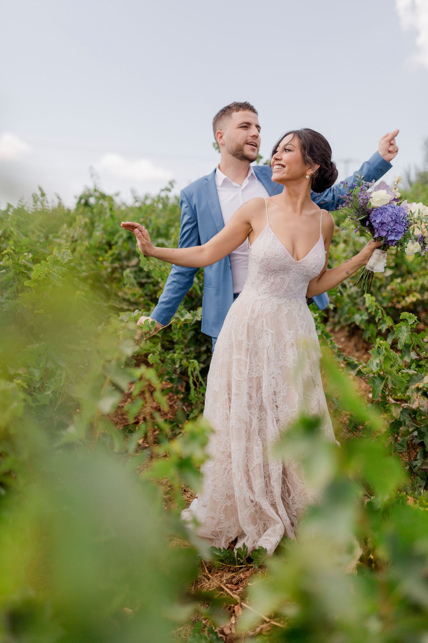 Wedding couple in vineyard, bride in lace dress holds hydrangea bouquet as groom in blue suit points.
