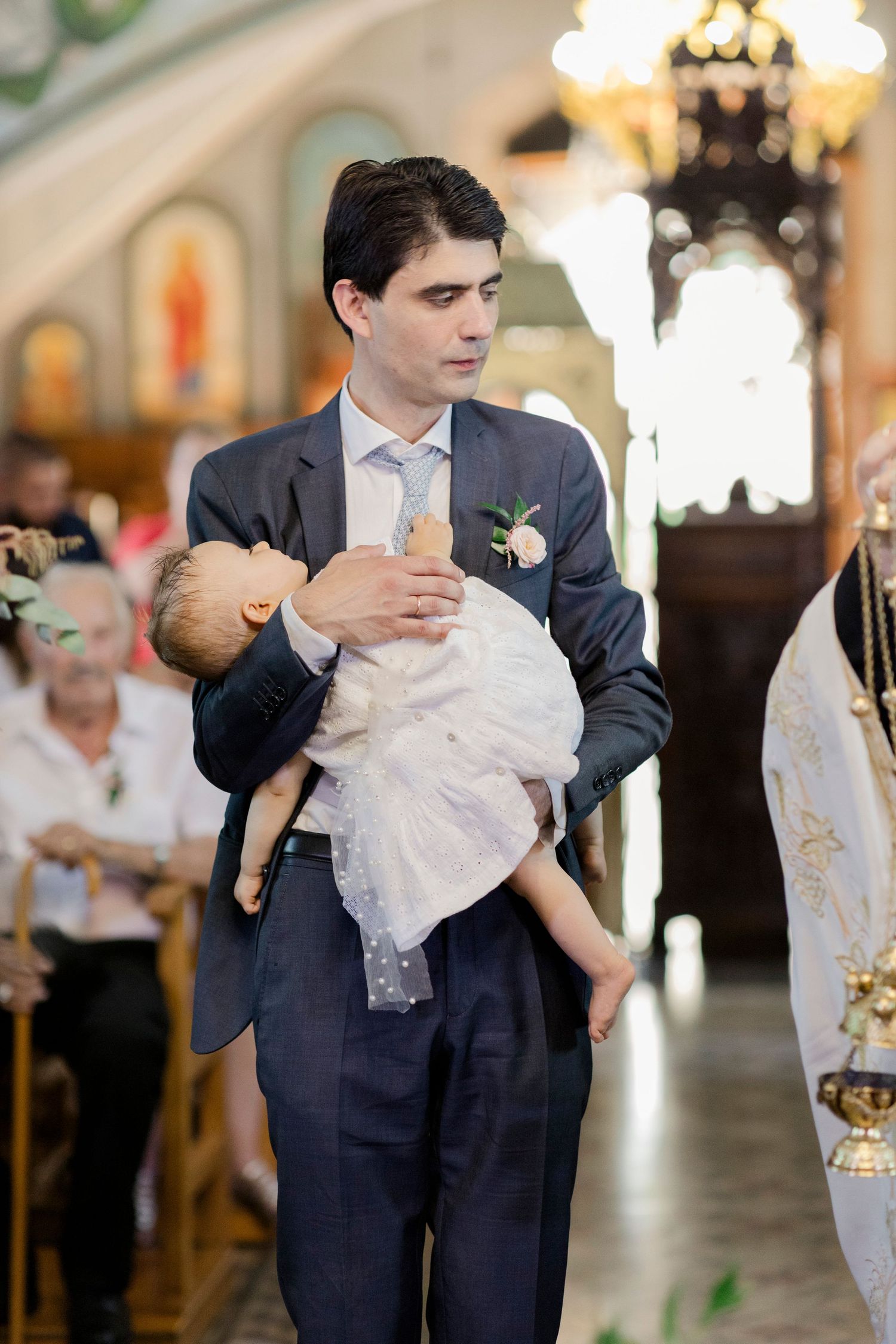 Man in suit holding baby in white christening gown during Orthodox church baptism ceremony.