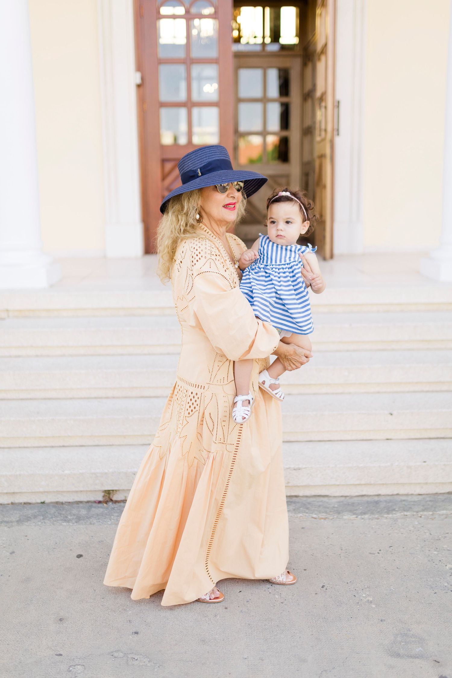 Woman in peach maxi dress and blue hat holds baby in striped outfit on white steps.