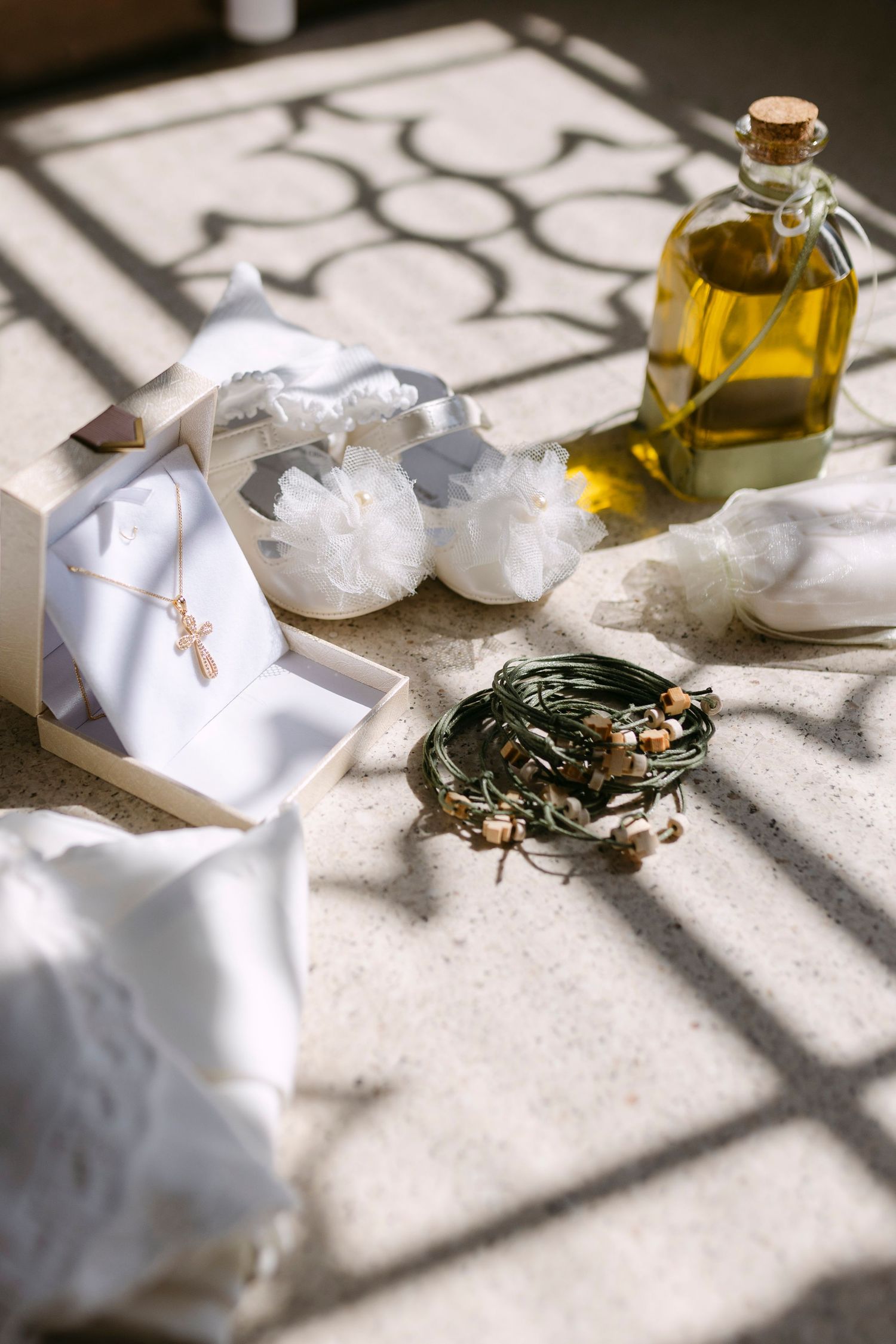 Greek Orthodox baptism items including white shoes, cross necklace, olive oil, and stefana crowns arranged in sunlight.